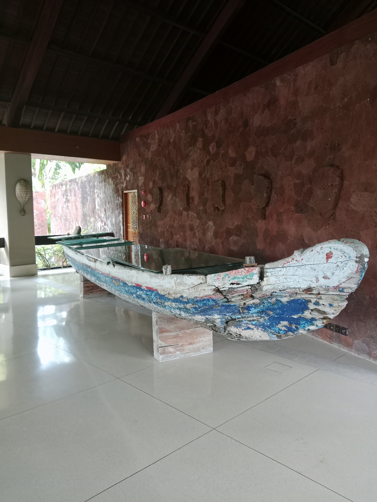 The image shows a large, weathered wooden boat that has been repurposed as a table. The boat is painted in a rustic style with white, blue, and hints of red. It is supported by two rectangular pedestals, and a glass top has been placed over the hull, transforming it into a functional table. The table is situated on a polished, light-colored tiled floor that reflects the light from the surrounding environment.
The background features a textured, reddish-brown wall made of irregular stones. Above the wall, a dark, sloped ceiling with visible wooden beams is present. To the left of the table, a large window or open doorway reveals lush greenery and what appears to be an outdoor area. A decorative woven object, resembling a tennis racket, hangs on a pillar to the far left. The overall setting suggests a space with an outdoor-indoor flow, possibly a reception area or a unique lounge in a resort or hotel. The environment is clean and well-lit, with natural light contributing to the ambiance. No people are visible in the image. The scene appears to be a still life, emphasizing the artistic repurposing of the boat. The location is identified as Kuta Selatan, Indonesia, which is known for its tropical climate and beach resorts, fitting the aesthetic of the image.