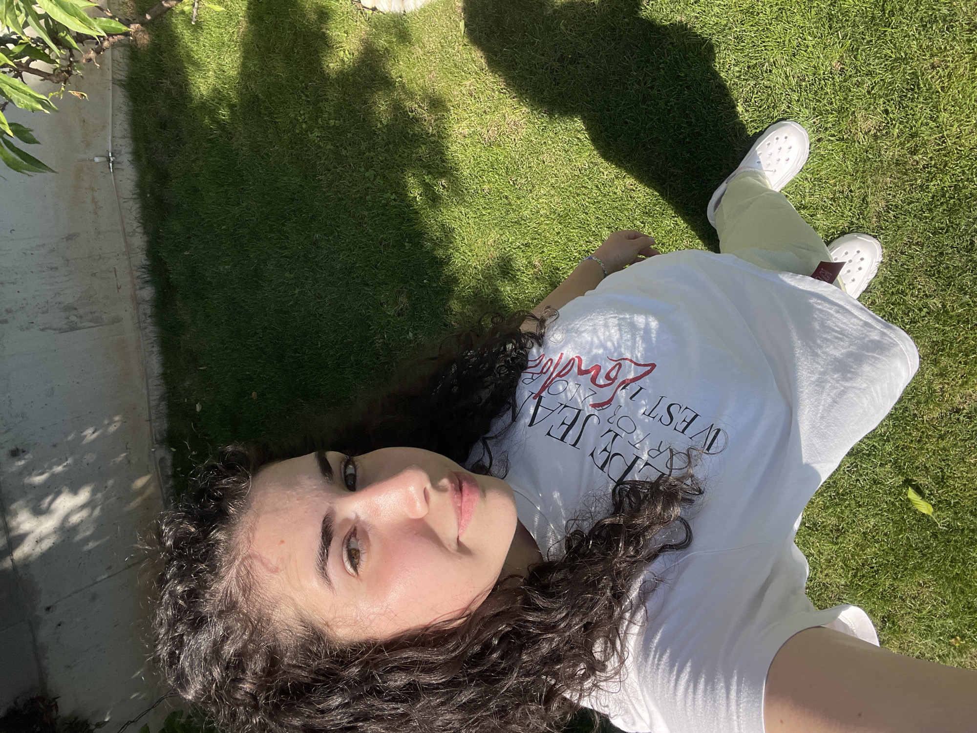 A young woman is lying on a grassy lawn, likely outdoors. She has dark, curly hair and fair skin. She is wearing a white t-shirt with black and red lettering and light-colored pants. Her feet are clad in white Crocs-style shoes. The scene is bathed in bright sunlight, casting shadows on the grass. A concrete wall can be seen in the background. The location is likely a park or garden in Sofia, Bulgaria, based on the vegetation, architectural style, and the style of the individual.