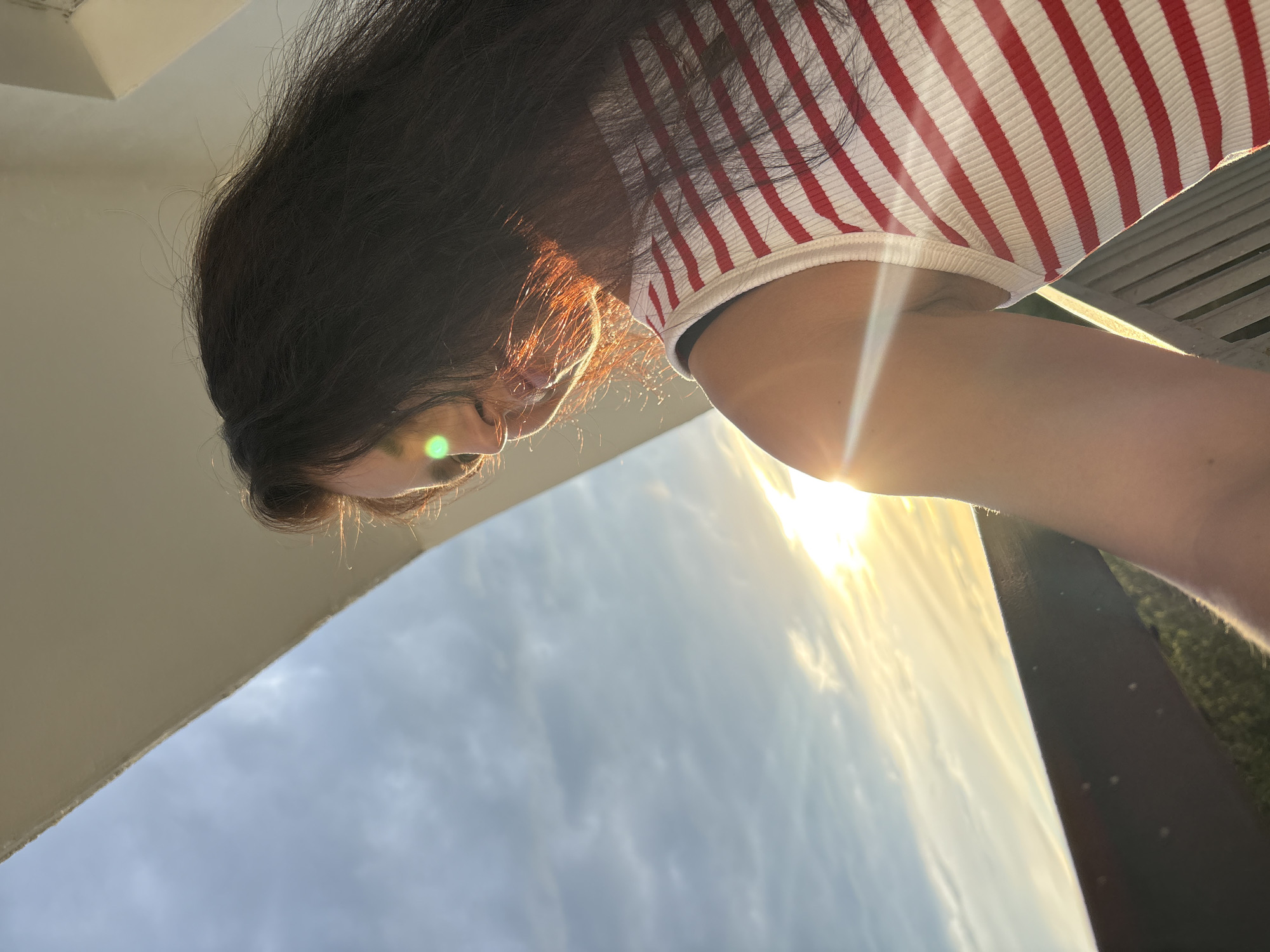 This is a close-up, rotated image of a young woman's head and shoulder, viewed from a low angle, possibly a selfie taken outdoors. She has long, dark hair that falls around her face and shoulders. She is wearing a white tank top with thin red stripes. The sun is shining brightly, creating a lens flare and casting a warm glow on her hair and skin. The background shows a bright sky with some clouds, suggesting it's daytime. A dark, slatted surface, possibly a bench or railing, is visible on the right side of the frame. The overall mood is relaxed and casual. The image appears to have been taken in Chaikovsky, Russia.