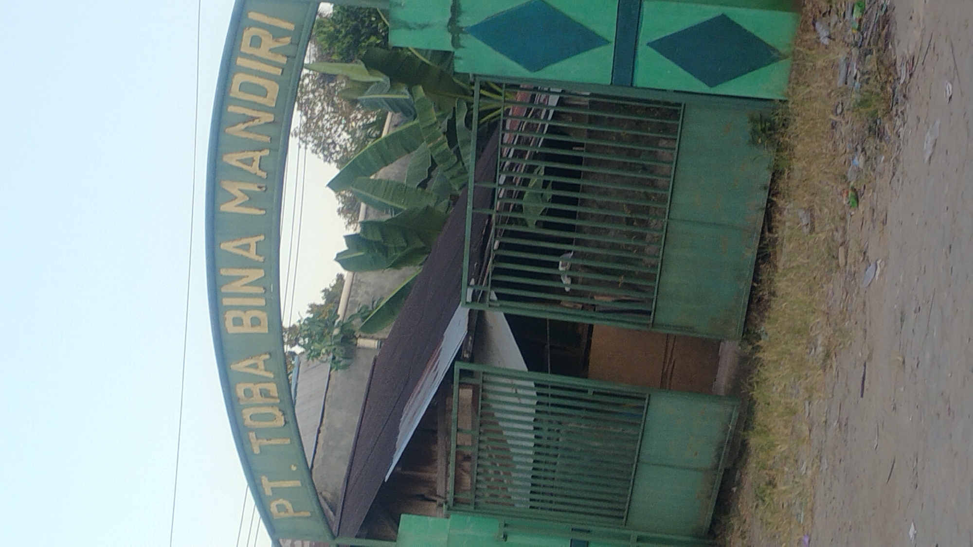 The image shows the entrance gate of PT. TOBA BINA MANDIRI in Samarinda, Indonesia. The gate is green with the company name in yellow letters on an arched sign above it. The gate is partially open, revealing a glimpse of a building and some vegetation behind it. The area in front of the gate is a dirt road with some dry grass and scattered debris. The overall scene suggests a commercial or industrial establishment in an outdoor setting.