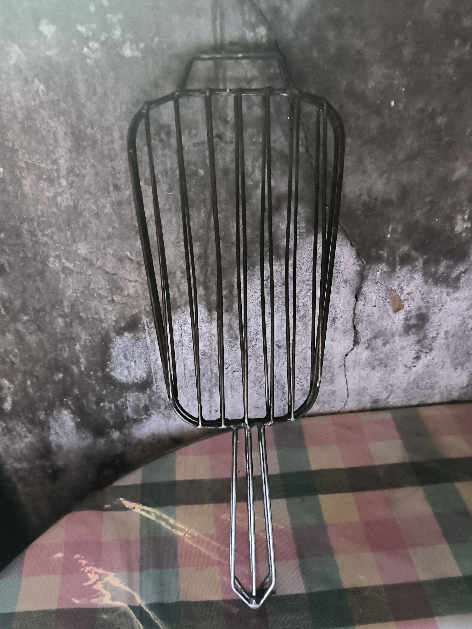 A metal grill basket stands vertically on a plaid surface in an indoor setting. The background is a textured, concrete wall. The location is Merbopuntong, Indonesia.