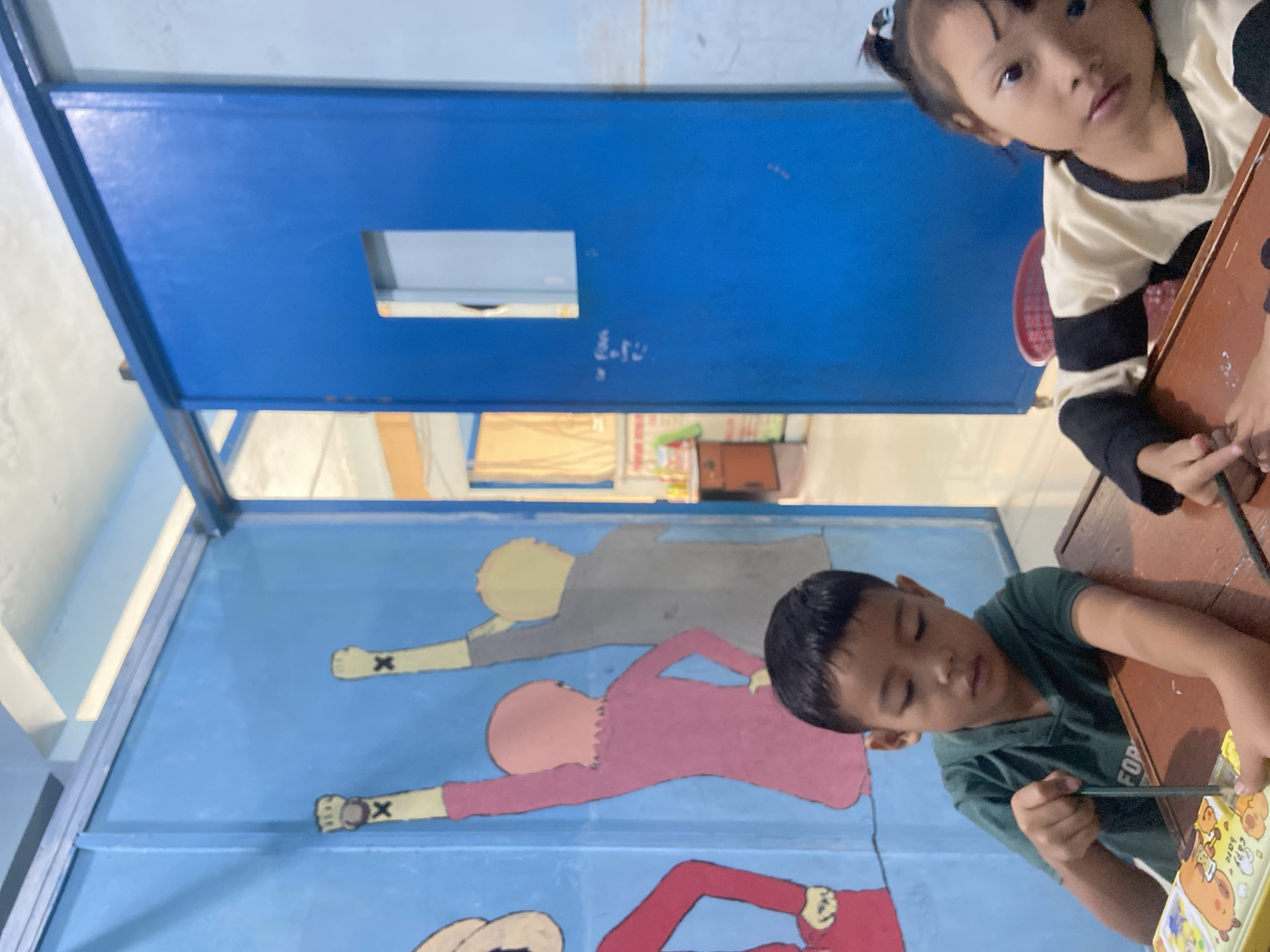 The image depicts an indoor scene, likely an educational or recreational facility, given the presence of children and a colorful mural on the wall. A young boy, wearing a dark green hooded shirt, is seated at a wooden table and appears to be engaged in an activity involving a yellow pencil case. He is holding a pencil and looking downwards. To his right, a young girl with her hair tied in a ponytail, wearing a light-colored shirt with black stripes, is also at the table, looking towards the camera.
The background features a bright blue sliding door with a rectangular cutout, and to the left, a mural painted on a blue wall depicts silhouetted figures with raised fists, each bearing an "X" mark. The environment suggests a room within a building, with elements like a ceiling and possibly other rooms visible through the doorway. There is a hint of a desk or counter in the background with some items on it, and a red plastic basket is visible behind the girl. The overall impression is of a casual, everyday setting with children participating in activities. The specific location within Tanjung Gading, Indonesia, cannot be definitively determined from the visual information alone.