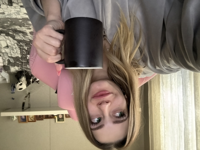 A young Caucasian female, with long blonde hair and green eyes, is depicted from the chest up in an indoor setting in Reutov, Russia. She wears a light grey hooded zip-up sweatshirt and is seated in a pink gaming-style chair. Her right hand holds a black cylindrical mug, exposing pink-painted fingernails. Her facial expression includes slightly puckered lips and eyes looking upwards and to her left, indicating a contemplative or posed glance. Behind her, a sheer, light-colored curtain covers a window on the left. A white floating shelf on the wall to the right displays several framed photographs and a small white figurine. Below the shelf, a black and white panda plush toy rests near a grey and white patterned duvet or blanket. The wall is a light, neutral color. The image appears to be a selfie.