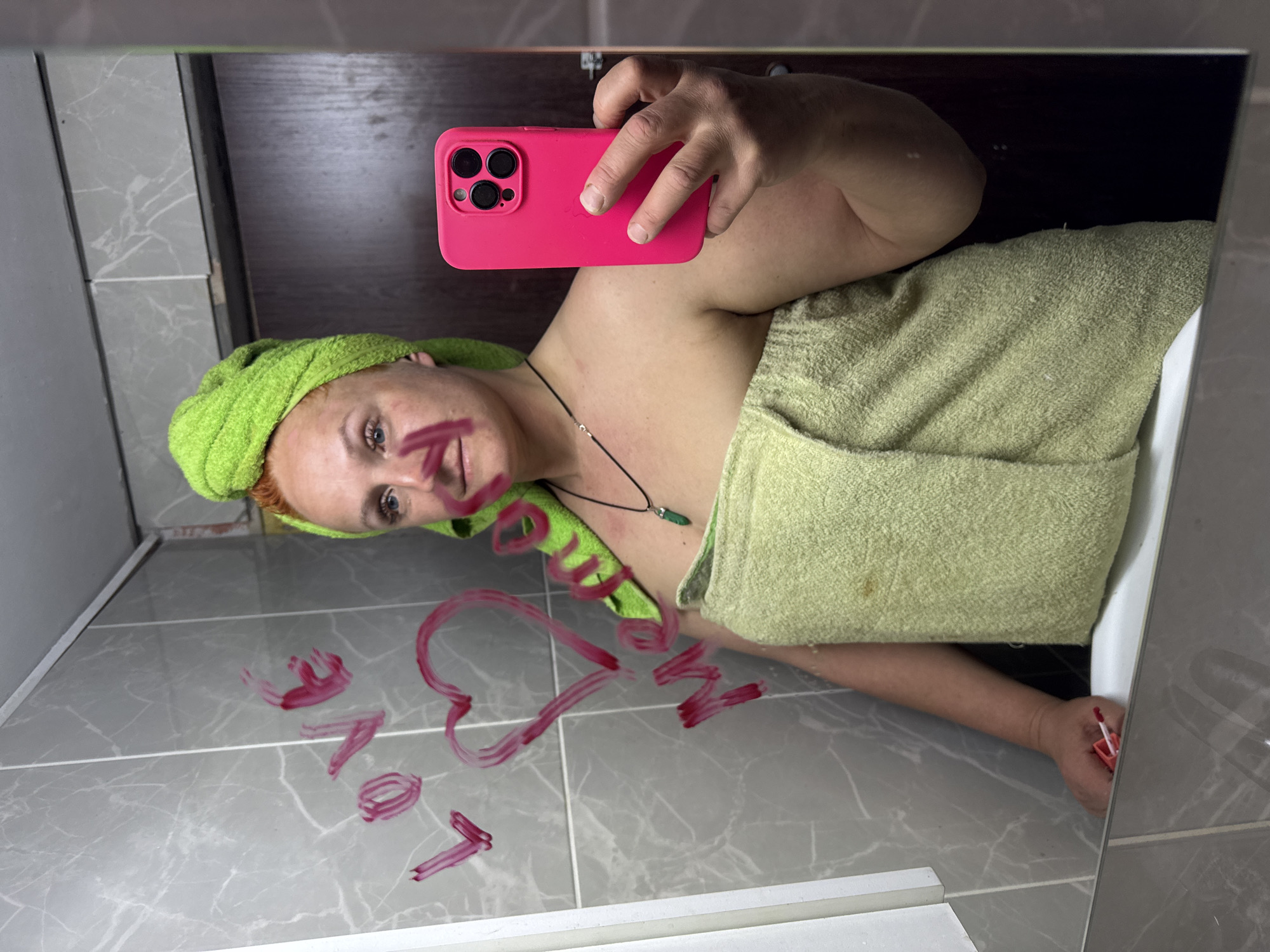 A person stands in a bathroom, taking a selfie in a mirror. The person is a fair-skinned individual with red hair, wearing a green towel wrapped around their head and body. They are holding a pink phone in their left hand. Written in red lipstick on the mirror is the word "love" along with a heart and two other symbols. The bathroom has grey tiled walls and a dark wooden door. The location is in Chaikovsky, Russia.