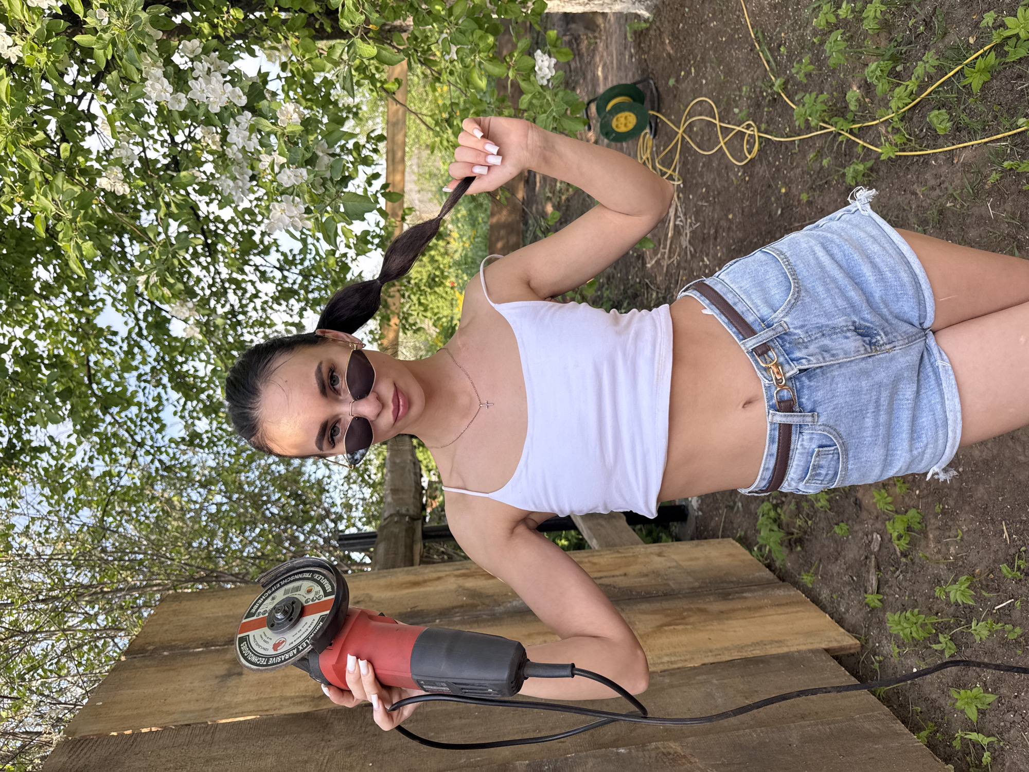 A woman stands outside in an area with scattered vegetation and a tree with white flowers. She is wearing sunglasses, a white tank top, and denim shorts with a brown belt. She holds a red and black angle grinder in her right hand and her braided ponytail in her left hand. She faces forward. A green hose reel sits in the background. She appears to be standing near wooden planks. The location is assumed to be the city of Chaikovsky, Russia.