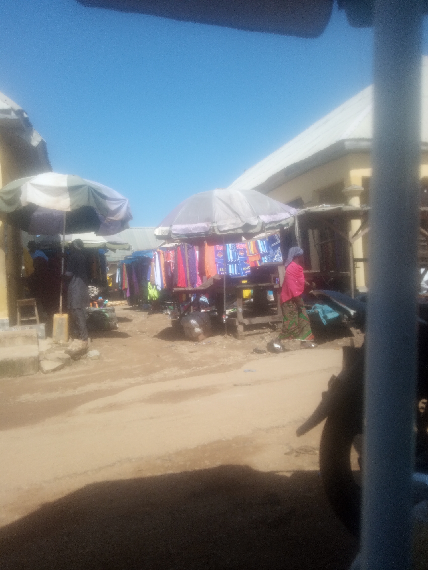 This image depicts an outdoor market scene in Serti, Nigeria, under a bright, clear blue sky, suggesting it is daytime with fair weather. The foreground is dominated by a dusty, unpaved ground with a large shadow cast from an unseen object, possibly a vehicle or structure.
In the mid-ground, several stalls are set up, covered by large umbrellas for shade. One stall prominently displays colorful fabrics and clothing items, hanging from a wooden structure. A person in a pink headscarf and patterned skirt is visible walking past this stall. Another stall to the left has a person in dark clothing standing behind a counter with more fabric displays. There are also bags and other merchandise visible on the ground near the stalls.
Further back, other market stalls and buildings with light-colored roofs are visible, extending into the background. The overall impression is of a bustling local market where goods are being sold under the open sky. The scene suggests a typical day in a Nigerian market, with vendors and customers engaged in commerce. There is no discernible text visible in the image.