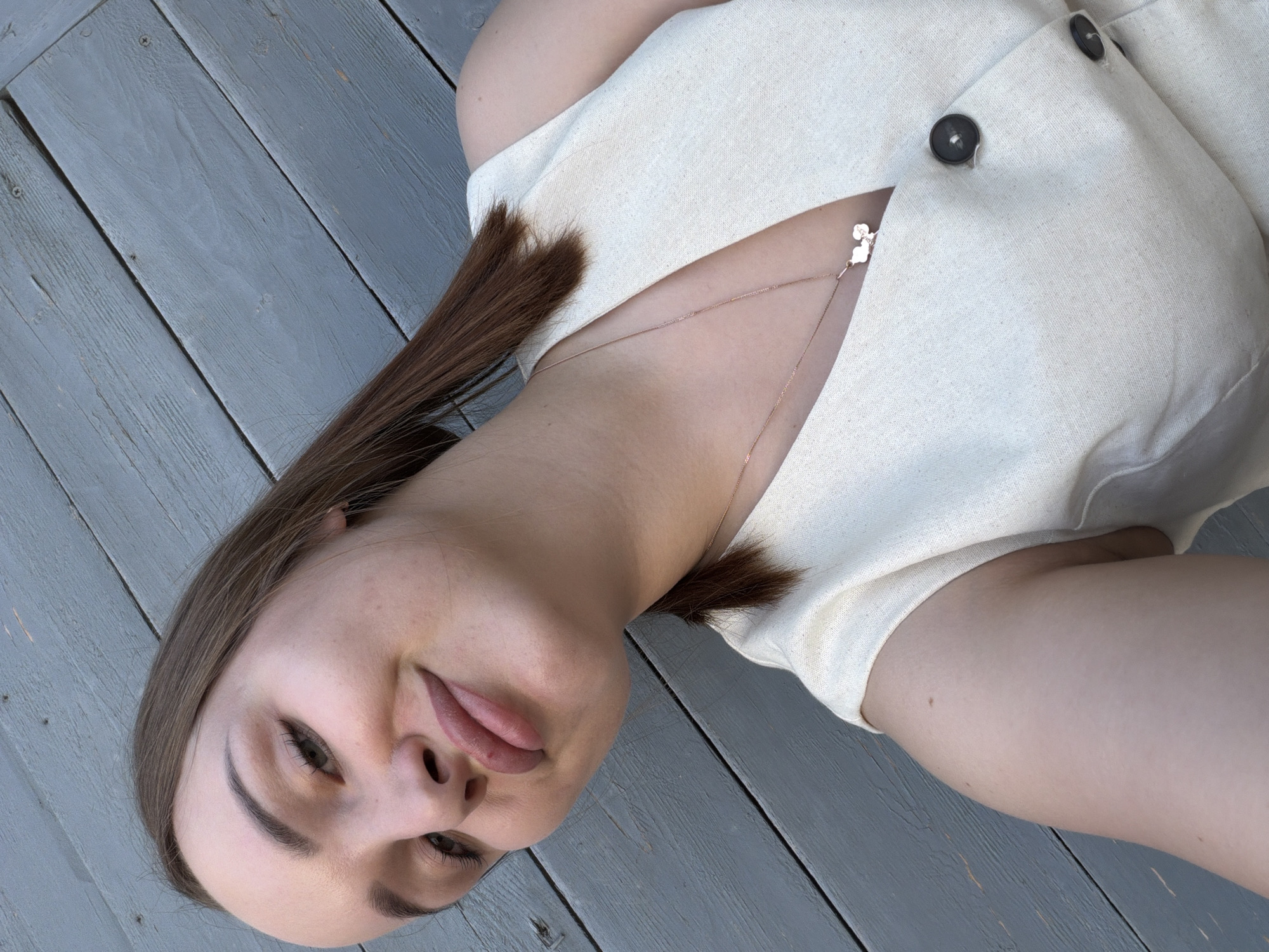 A woman with fair skin and light brown hair is lying on a gray wooden surface. She is wearing a light-colored dress with buttons. A delicate necklace is visible around her neck. The location is Chaikovsky, Russia.