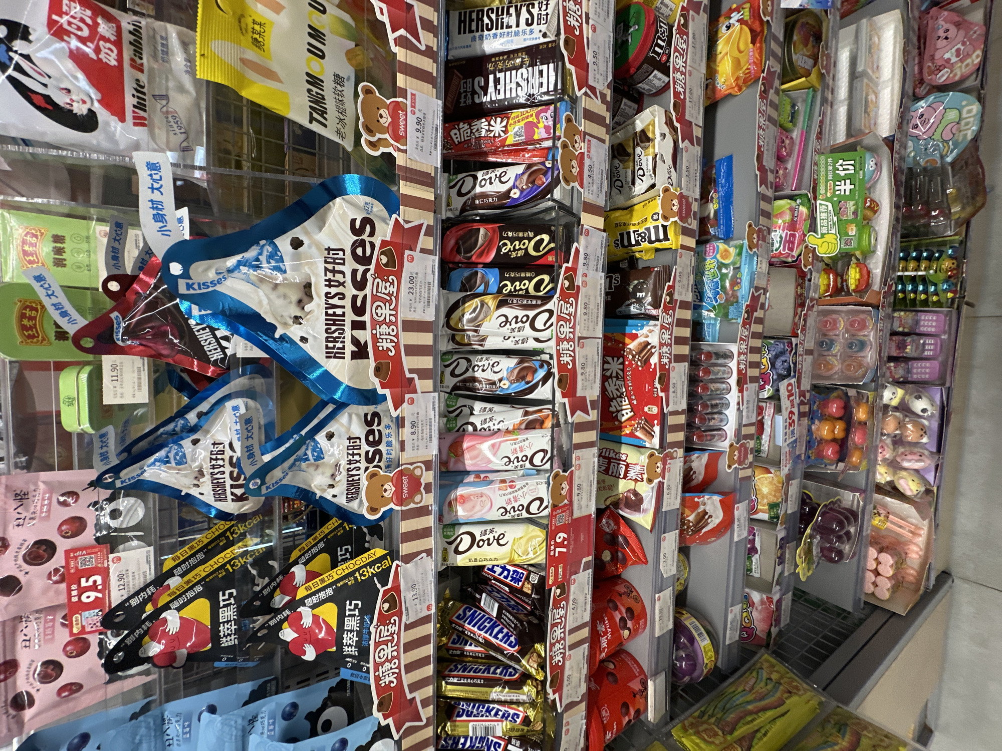 The image shows a well-stocked candy display in a retail setting, likely a convenience store, located in the city of Chongqing High-tech Zone, China. The merchandise includes a variety of candies and chocolates, such as Hershey's Kisses, Dove chocolates, Snickers bars, M&M's, and local Chinese brands like White Rabbit candy and Tang Mumu. Prices are prominently displayed in Chinese Yuan (¥), indicating the local market. The shelving is wire grid, with merchandise hanging from hooks or stacked on shelves with price tags attached to dividers. The display also includes small novelty items like erasers or stationery alongside the candies.