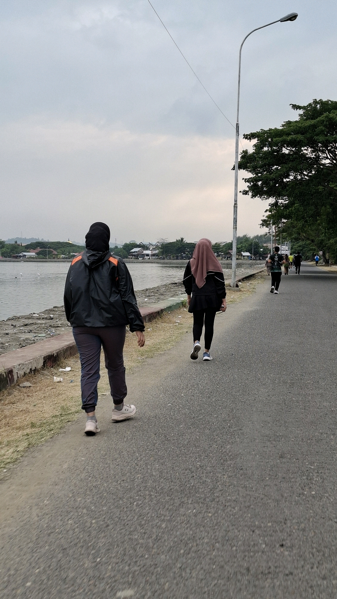 The image shows a scene at the Lhokseumawe Reservoir in Lhokseumawe, Indonesia. Two women, dressed in modest athletic wear with headscarves, are walking along a paved path next to the reservoir. The woman in the foreground wears a black jacket with orange accents and grey pants, while the woman behind her wears a black outfit and a pink headscarf. Several other individuals are visible walking or running further down the path, suggesting a recreational area. The reservoir's water is visible to the left, with some debris along its edge. In the background, a tree-lined area and distant buildings indicate a populated urban or suburban environment. A lamppost stands on the right side of the path, and a power line stretches across the sky, which is overcast. The overall atmosphere suggests a calm, outdoor activity session.