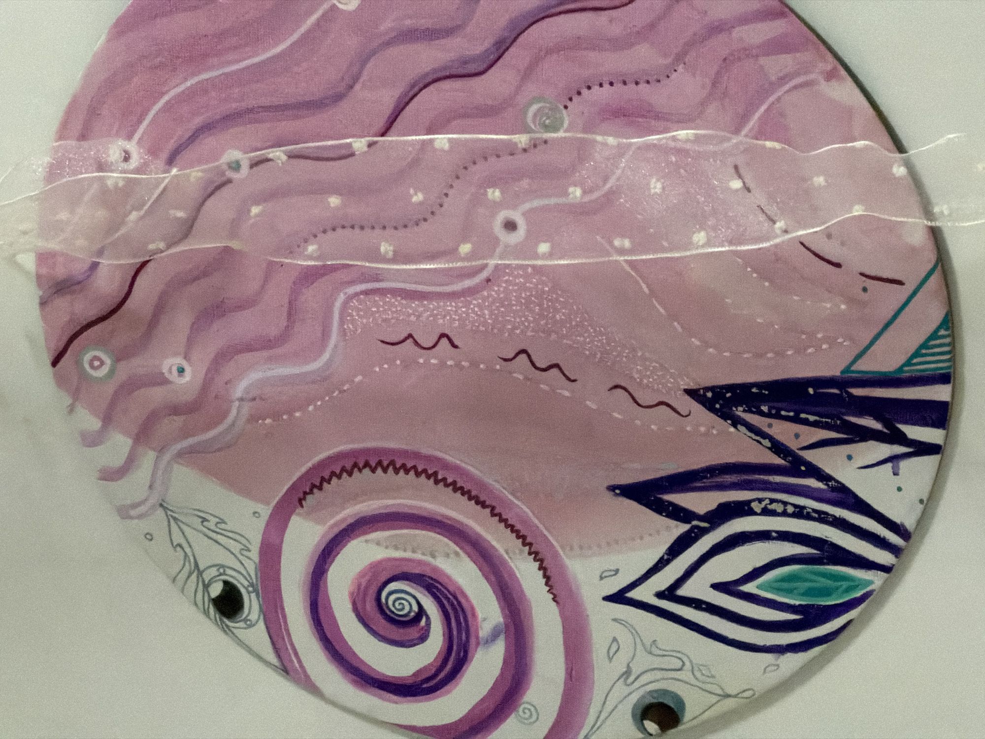 The image is a close-up of a round, hand-painted object, likely a decorative piece or part of a larger artwork. The primary colors are various shades of pink, with accents of purple, blue, and teal. The painting features abstract swirling patterns, wavy lines, and what appear to be stylized floral or flame-like shapes along the bottom edge. There is also a drawing of an eye on the left side of the object. A sheer, translucent ribbon with a subtle dotted pattern is draped vertically across the right half of the object, obscuring some of the painted details. The background is a plain, light-colored surface, likely a wall or table, with a subtle shadow cast on the right side. The image is a still life, and no people or specific locations are visible. This image is not from Moscow, Russia.