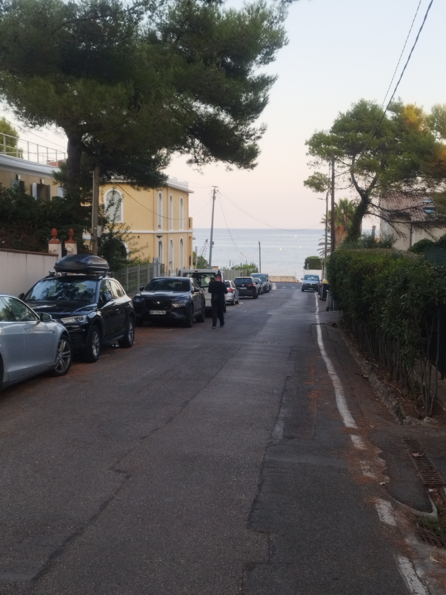 The image depicts a street scene in Antibes, France, leading towards the sea. Several cars are parked on the left side of the street, including a black Audi SUV with a roof box and a black Jaguar sedan. A person in dark clothing is walking away from the camera on the right side of the street. The street is paved with asphalt and has a white line marking the edge. Trees and bushes line the right side of the street, and residential buildings are visible on the left and in the background. The sky is clear and the sea is visible in the distance. The lighting suggests it is either early morning or late afternoon.