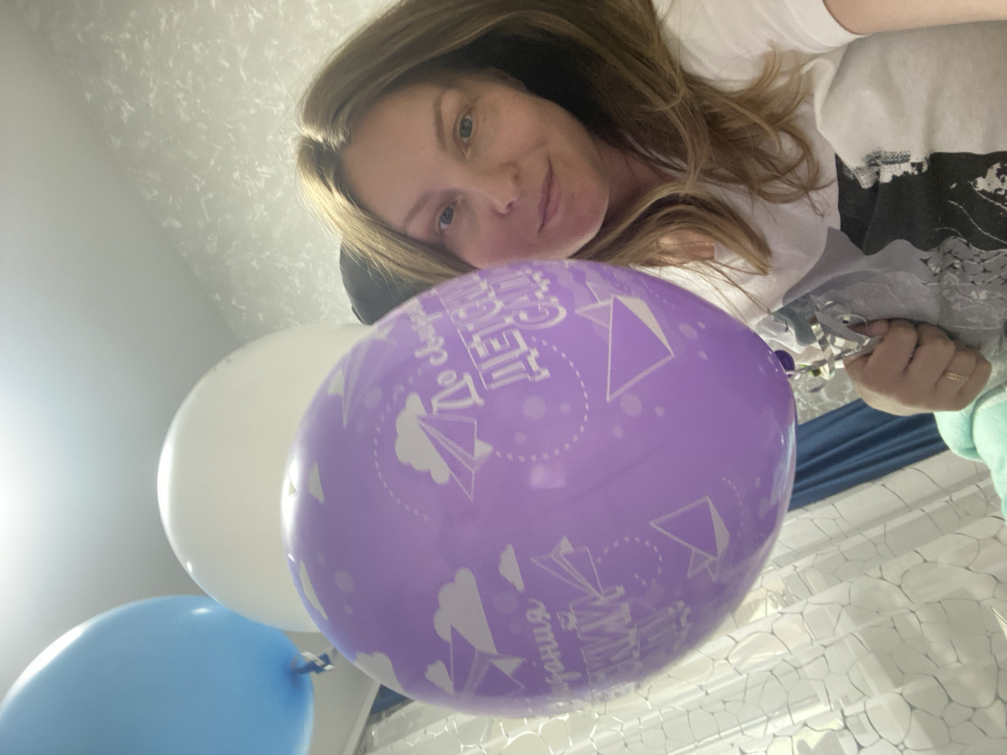 A woman is taking a selfie with balloons. The balloons are blue, white, and purple and have a text on them in the Russian language. The woman is wearing a white t-shirt. The background is blurry, but it appears to be a room with a window and a curtain. The image was taken in Noviy Urengoy, Russia.