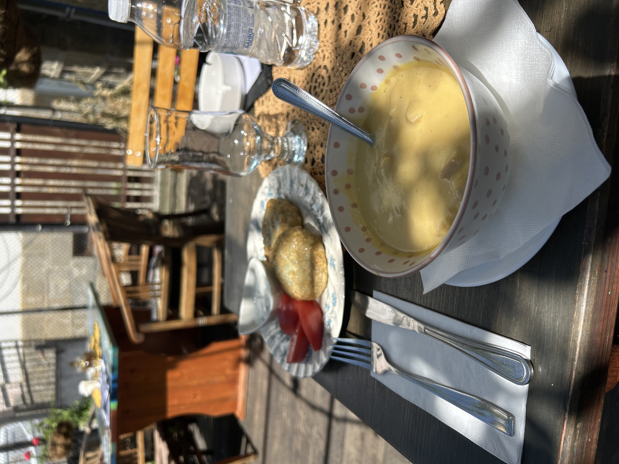 The image shows a table setting, likely in an outdoor area in Troyan, Bulgaria. The table is set with a bowl of soup, a plate with what appears to be fried cheese, tomato slices, and a small dipping sauce, a glass, a bottle of water, silverware wrapped in a white napkin, and a knitted placemat. The table appears to be made of dark wood. Behind the table, one can see a wooden chair and a glimpse of the surrounding environment, including buildings and greenery, indicating an outdoor restaurant or cafe setting in Troyan. The lighting suggests it is daytime. There are no people visible in the image.
