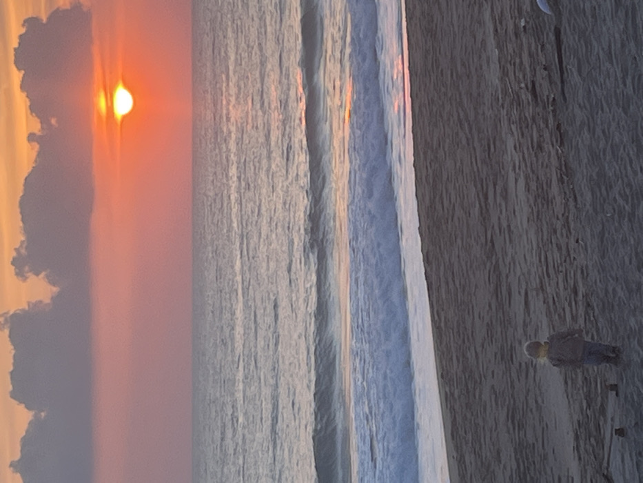 The image depicts a sunset over the Mediterranean Sea from a sandy beach in Alanya, Turkey, likely a westward-facing stretch such as Cleopatra Beach. The sky exhibits a gradient of vibrant orange and pink hues near the horizon, transitioning to lighter pink and grey above, with several dark, irregular cumulus clouds prominent in the upper-left area. The sun, appearing as a bright, flattened orange-yellow orb, is positioned just above the sea horizon, casting a shimmering light path across the water's surface. The sea itself is a muted grey-blue, with multiple waves breaking into white foam along the shoreline. In the foreground, a single small child, dressed in a grey long-sleeved top and blue trousers, stands on the dark grey sand, facing away from the camera and looking towards the sea. Scattered dark, unidentifiable debris is visible on the sand to the child's right. No other individuals or significant structures are discernible.