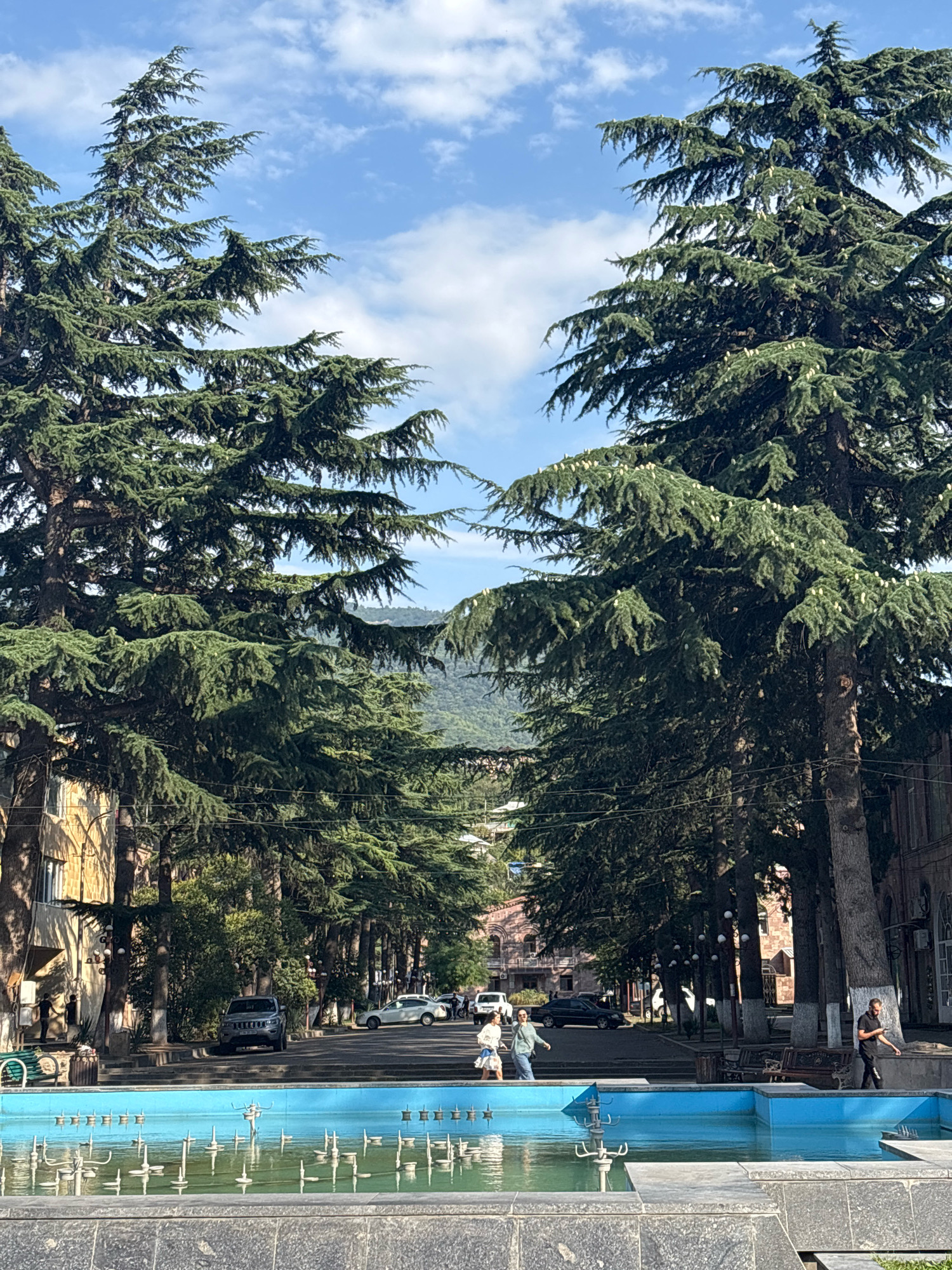 This image captures a street scene in Ijevan, Armenia, dominated by large, mature trees lining both sides of the road, creating a natural archway. In the foreground, a bright blue, empty fountain with its intricate metal spouts is visible. Beyond the fountain, a street stretches into the distance, with a line of cars parked and driving along it, including a silver SUV and a white sedan. Several people are walking along the sidewalk and street. Two women are walking together on the left side of the street, and a man is walking on the right side, looking at his phone. Buildings with traditional architecture, some appearing to be historic, flank the street. In the background, rolling green hills are visible under a partly cloudy sky, suggesting a pleasant day. The overall atmosphere is one of a peaceful, tree-lined urban environment with elements of everyday life unfolding.