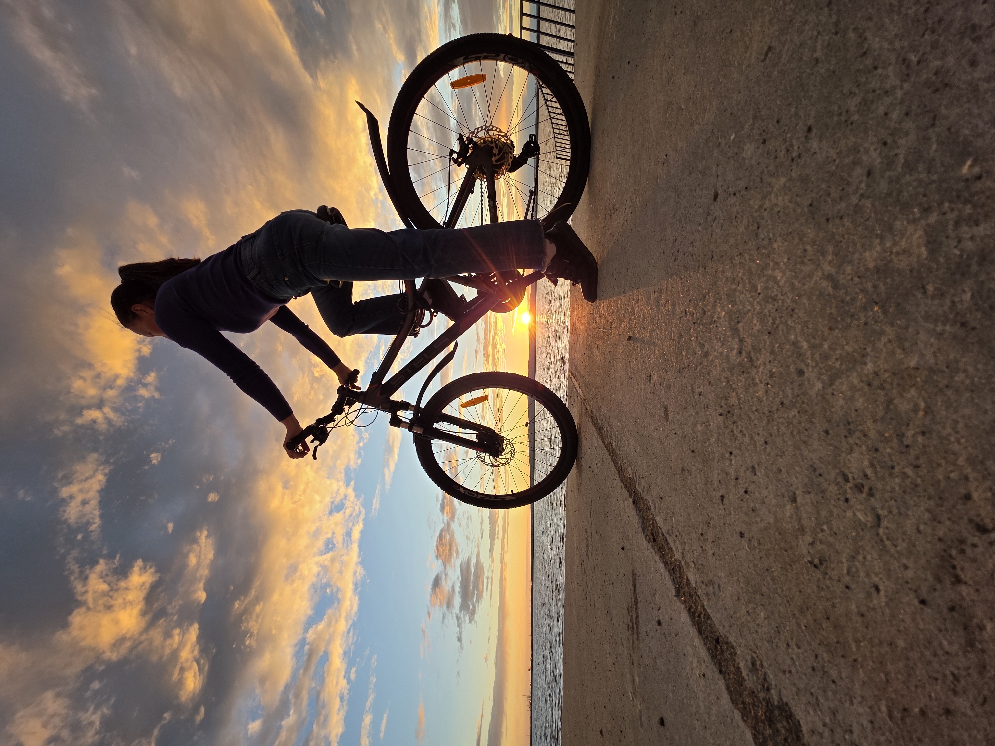 A silhouette of a woman on a bicycle is visible against a dramatic sunset sky. She is wearing a dark long-sleeved shirt and blue jeans, with her hair pulled back in a ponytail. The bicycle is dark-colored with visible front and rear suspension and disc brakes. The woman is positioned on a paved surface next to a body of water, likely the Kama River, given the location of Chaikovsky, Russia. The sun is low on the horizon, casting a warm glow and creating long shadows. The sky is filled with colorful clouds in shades of orange, yellow, and blue. The foreground shows the textured pavement with a faint tire track.