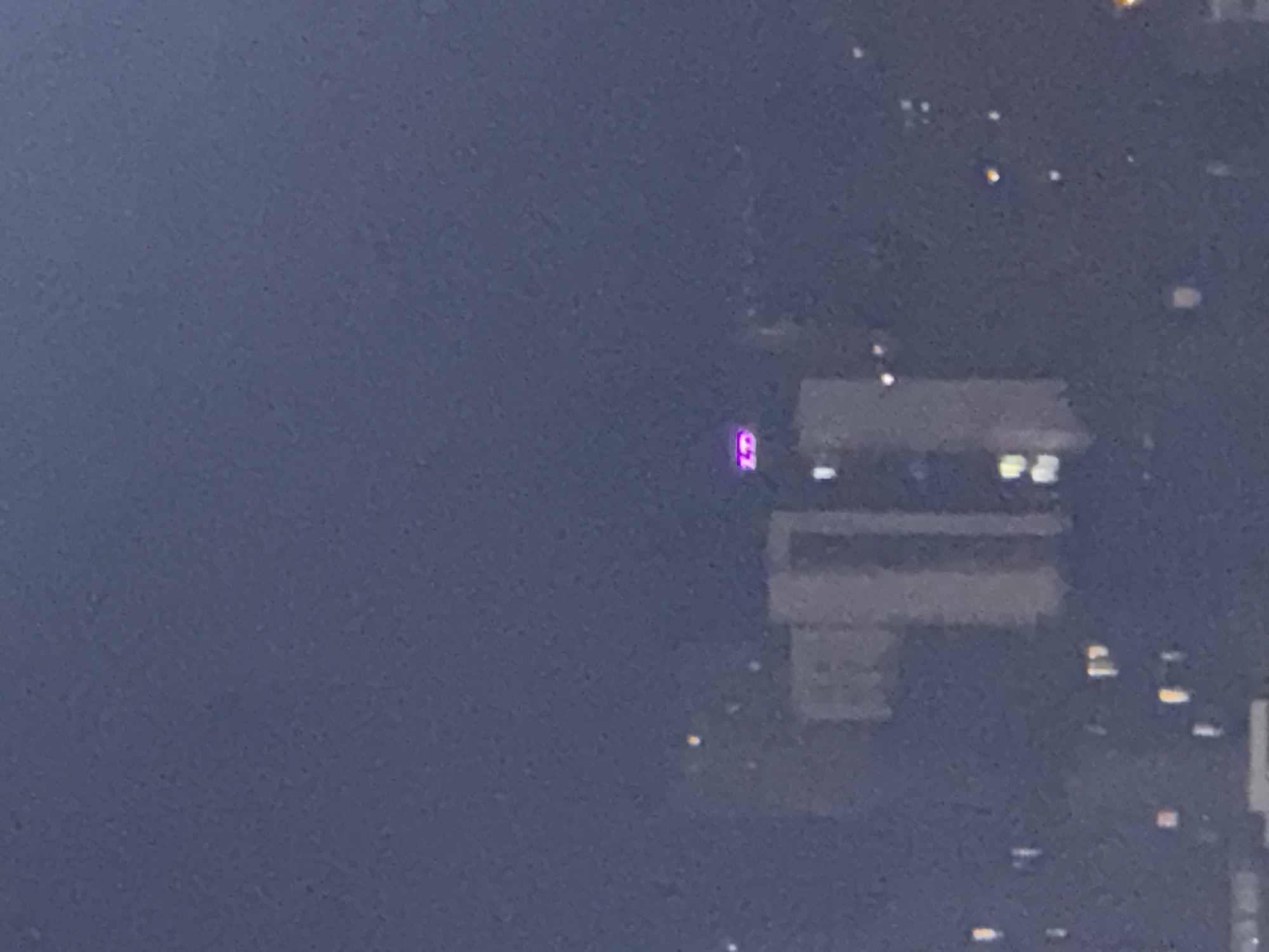 The image shows a nighttime view of a cityscape, likely Tbilisi, Georgia, given the presence of a building with illuminated signage. In the mid-ground, a tall, modern building is visible, its upper section adorned with bright pink neon lettering that appears to spell out "LD". The rest of the building is dimly lit, with some windows glowing yellow. The surrounding environment is dark, with scattered points of light from other buildings and streetlights, creating a blurred, atmospheric effect. The overall impression is one of urban activity at night, with the neon sign serving as a focal point.
