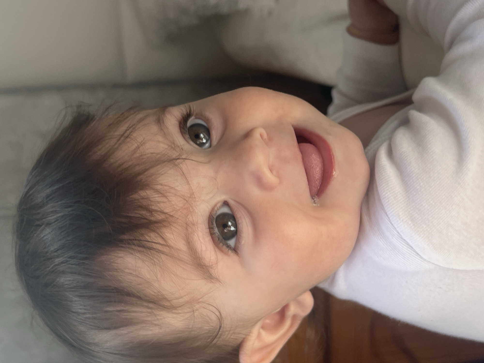 The image presents a close-up, rotated portrait of an infant, with the baby's head positioned on the left side of the frame and the body extending towards the right. The baby has a fair complexion, captivating dark brown eyes, and fine, dark hair. A curious and wide-eyed expression is directed towards the viewer, with the tongue slightly peeking out from the mouth, accompanied by a small amount of moisture or drool on the lower lip. The infant is dressed in a white, ribbed long-sleeved top. The background is softly blurred and largely indistinct, suggesting an indoor setting, possibly a room with light-colored furniture or walls. The lighting is soft and natural, indicating that the photo was taken during the day. No text is visible in the media file. This moment was captured in Elena, Bulgaria.