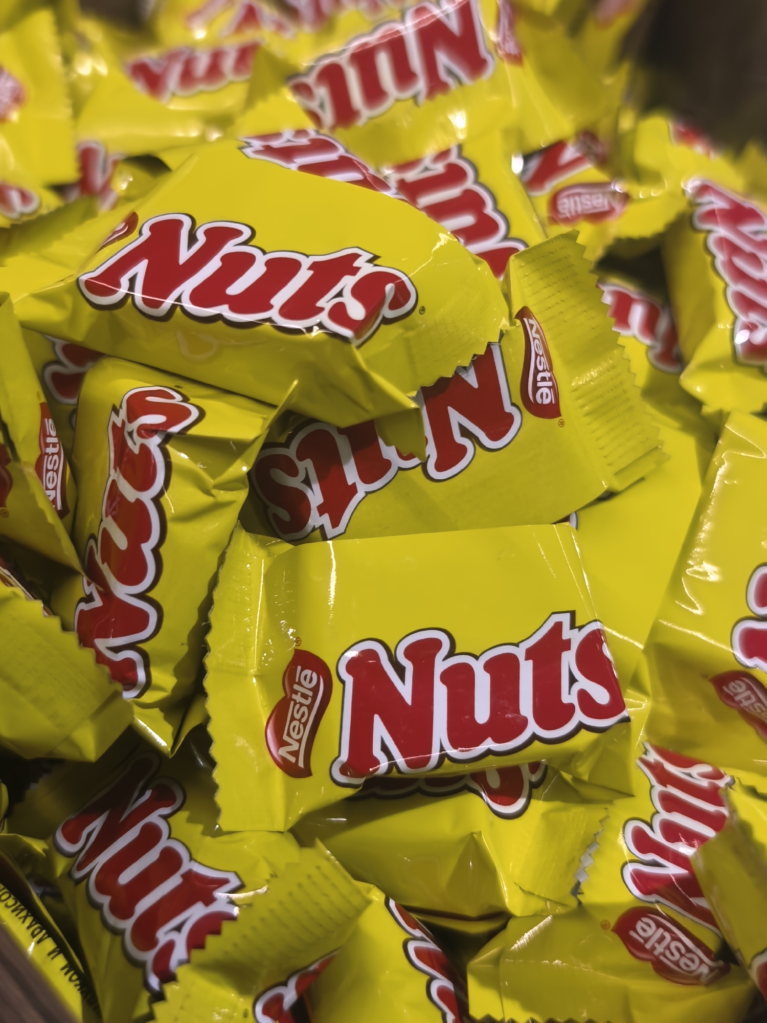 A close-up, overhead shot displays a pile of individually wrapped Nestlé Nuts candy bars. The wrappers are bright yellow with the red and white "Nuts" logo prominently featured in a stylized font. The logo is visible on multiple candy bars, sometimes oriented horizontally and sometimes vertically, depending on how the wrappers are positioned. The Nestlé logo is also visible on some of the wrappers in its characteristic red oval shape. The candy bars are stacked unevenly, creating a textured, overlapping pattern. The lighting is bright, suggesting an indoor setting with artificial light, or possibly a bright, sunny day if outdoors. There are no people visible in the image. The focus is solely on the product, hinting at a commercial display or a bulk purchase of the candy. Some wrappers show slight crinkling and creasing, indicating they are standard retail packaging. The overall impression is one of abundance and accessibility of the candy.