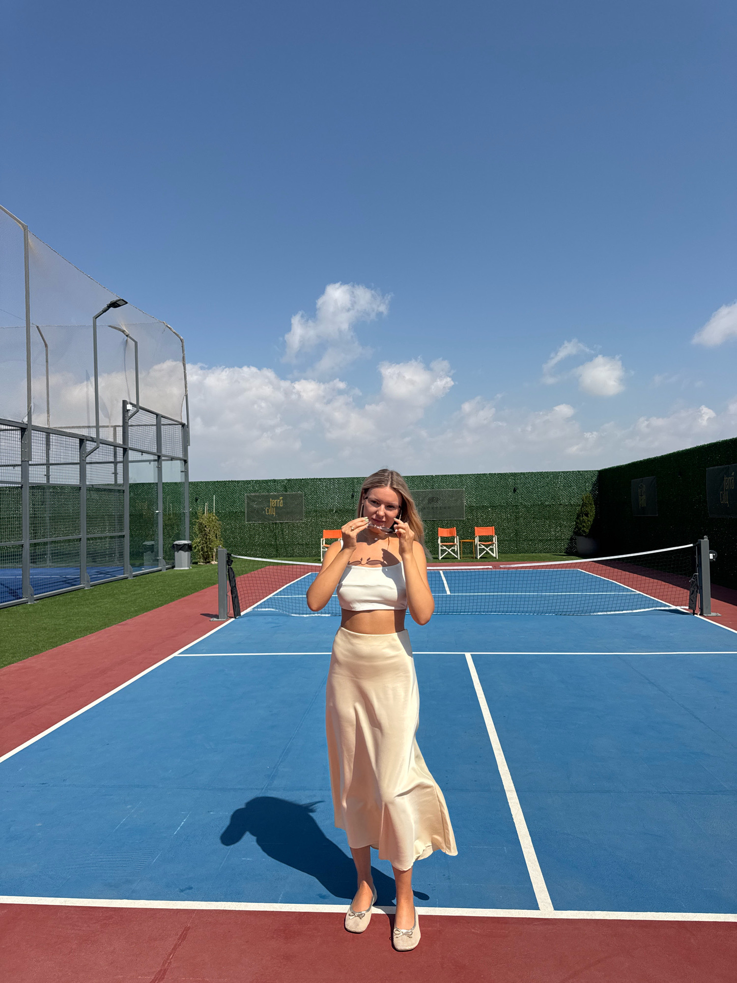 A young woman stands on a blue and red tennis court under a clear blue sky with scattered white clouds. She is wearing a white crop top and a long, flowing cream-colored skirt, and she is holding a pair of sunglasses. The court is surrounded by green netting and artificial turf. In the background, there are orange chairs and a logo that reads "Terra City". The scene is set outdoors on a sunny day. The location is a tennis court in Antalya, Turkey.