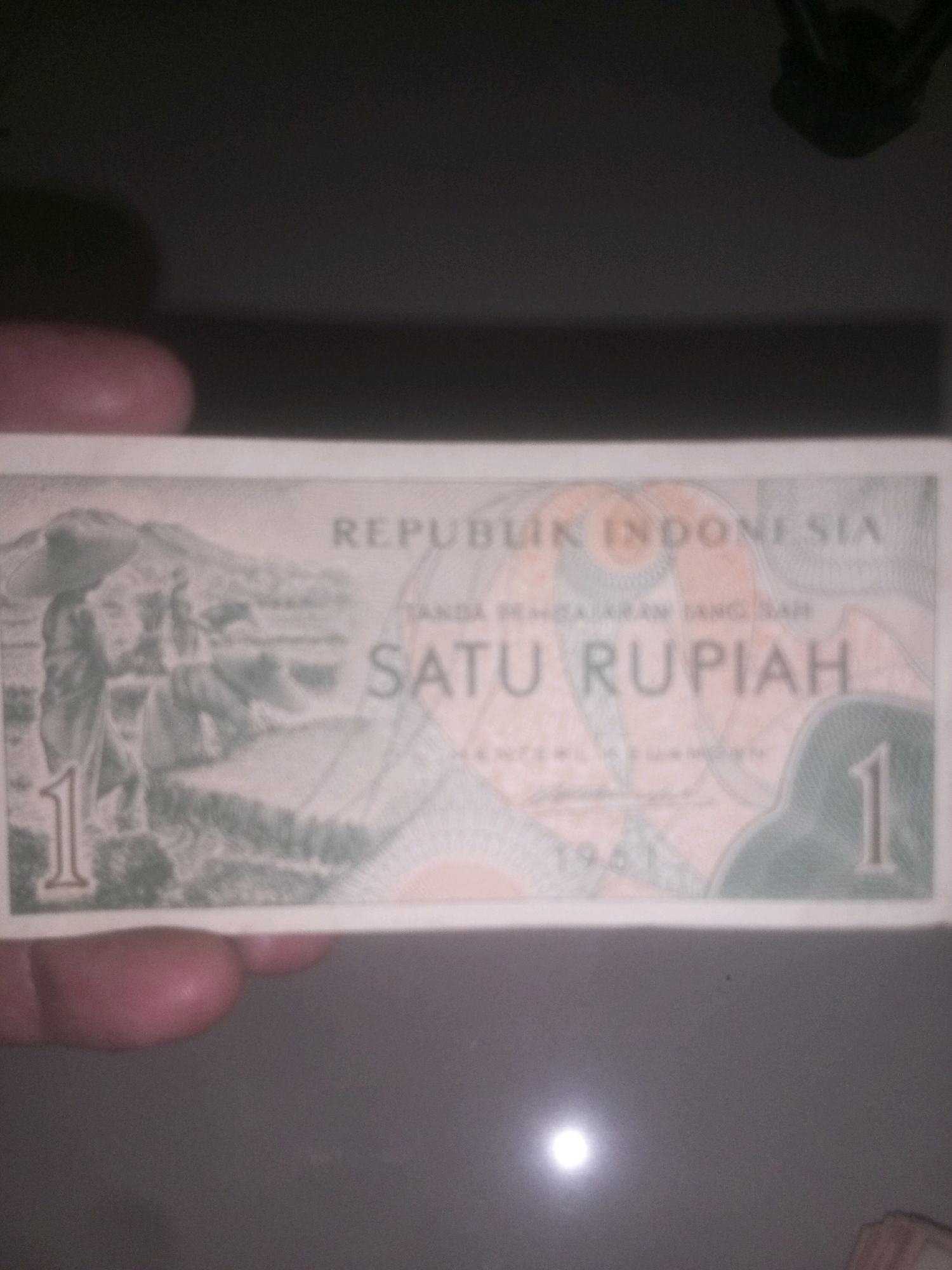 The image shows a close-up of a 1961 Indonesian one rupiah banknote. The banknote is light green and features an agricultural scene with farmers working in a field. The text on the banknote reads "REPUBLIK INDONESIA" at the top and "SATU RUPIAH" in the center. The numeral "1" appears prominently on the left and right sides of the banknote. The banknote is held by a person whose fingers are visible on the bottom left of the image. The background is dark and out of focus, suggesting the photo was taken indoors with poor lighting conditions. There is no visible indication of the location being the city of Kediri City, Indonesia.