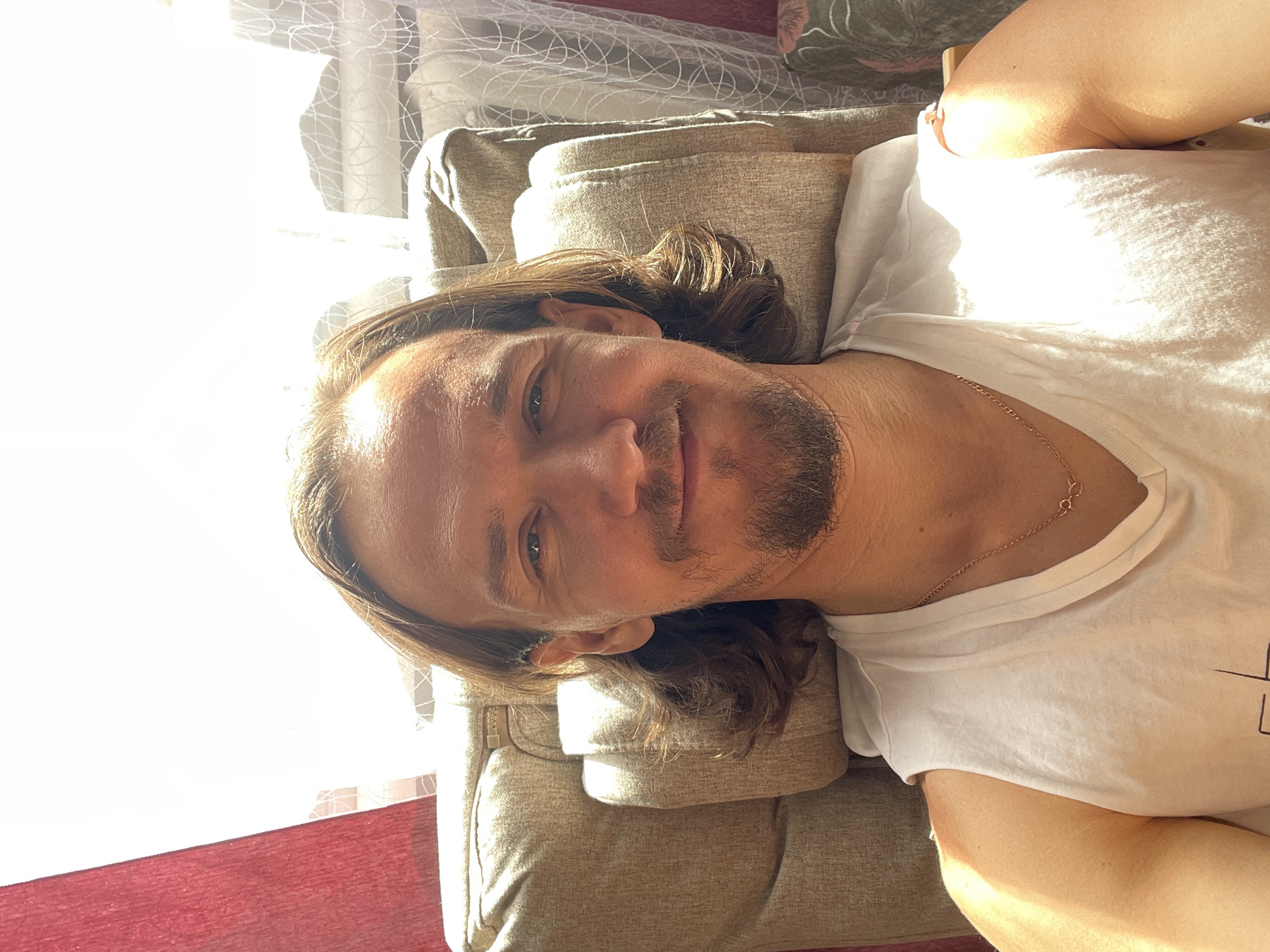 A man with long brown hair and a beard is lying on a light brown couch. He is wearing a white t-shirt and a gold necklace. The man is looking at the camera and smiling slightly. There is a window with white curtains in the background. A red surface is visible near the window. The image was taken in the city of Перевозное, Russia.