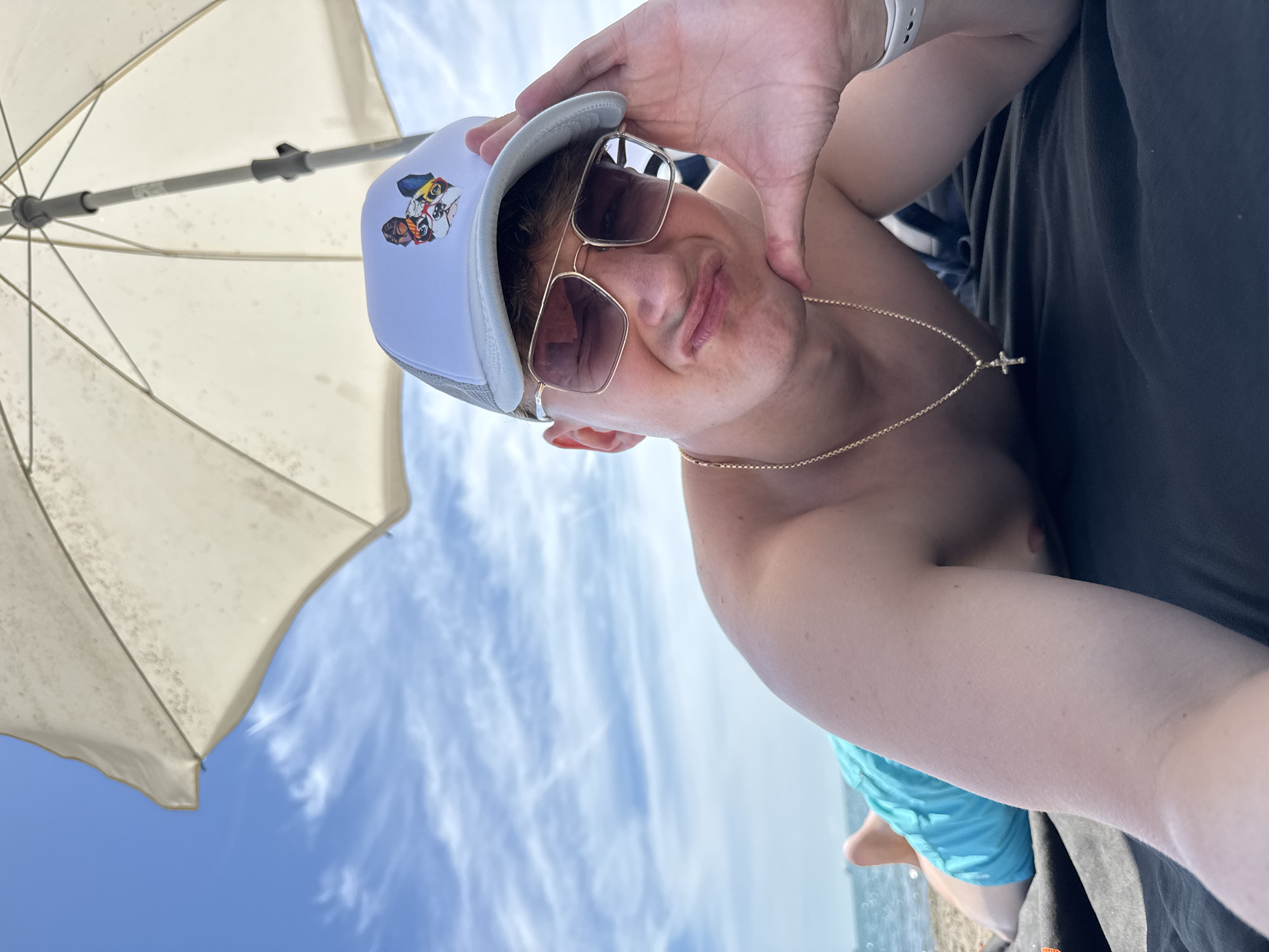 A young man wearing a white trucker hat with a dog graphic, gold-rimmed sunglasses, and a gold chain with a cross pendant lies on a dark beach towel under a white beach umbrella. He is shirtless and wearing turquoise swim trunks. His left hand is propped on his cheek, and he is making a pouty face at the camera. The background shows a blue sky with white clouds and the sea. The location is the beach in Burgas, Bulgaria.