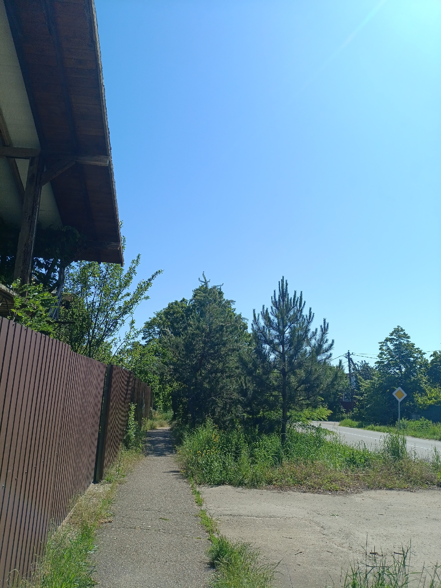 This photograph captures a bright, sunny outdoor scene in Goryachiy Klyuch, Russia, likely during mid-day. The clear blue sky dominates the upper portion, featuring a visible sun flare in the top right corner.
On the left side of the image, the underside of a brown wooden roof structure is visible, extending over a brown corrugated metal fence. This fence runs along a paved path or sidewalk that stretches into the distance. The path is bordered by green grass and various wild plants, which appear somewhat overgrown.
To the right of the path and vegetation, a paved road curves gently into the distance. Along the roadside, there are more dense green trees, including conifers and deciduous types, forming a natural backdrop. A yellow diamond-shaped road sign, typically indicating priority, is visible on the right side of the road, partially obscured by foliage. Utility poles and wires can be faintly seen among the distant trees on the right.
No people are visible, and no specific activity is taking place; the image presents a static landscape view, combining man-made structures with a lush natural environment under clear skies. There is no visible text.