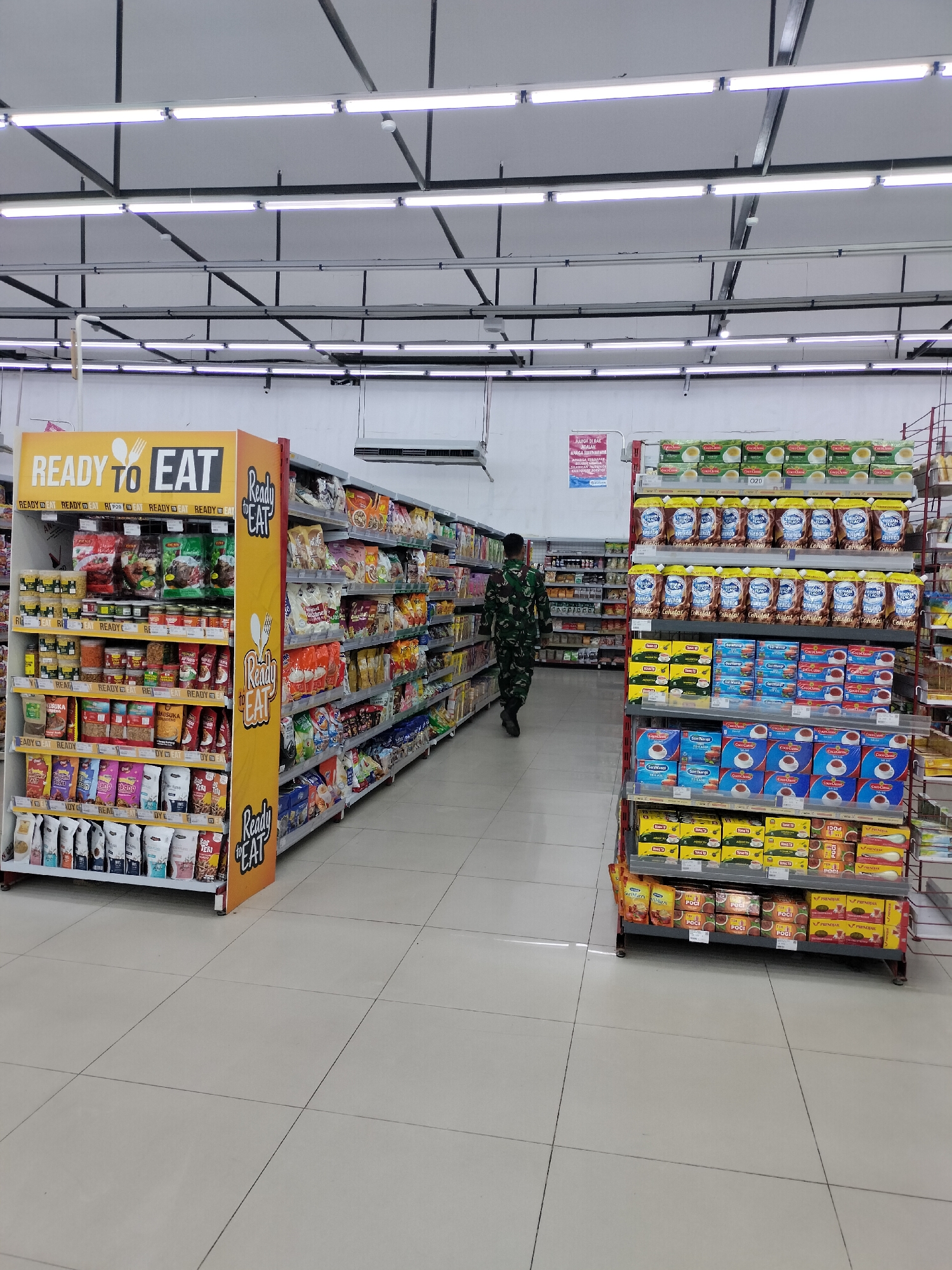 The image shows the interior of a supermarket in Langsa, Indonesia. In the foreground, a yellow display rack with the "READY TO EAT" logo is filled with various food products, including packets of snacks and instant meals. To the right of this display, a person in a camouflage military uniform is walking away from the camera down an aisle lined with shelves stocked with packaged beverages, likely milk and coffee, and other food items. The store is well-lit with fluorescent lights and has tiled flooring. The shelves in the background are packed with more merchandise, suggesting a busy retail environment.