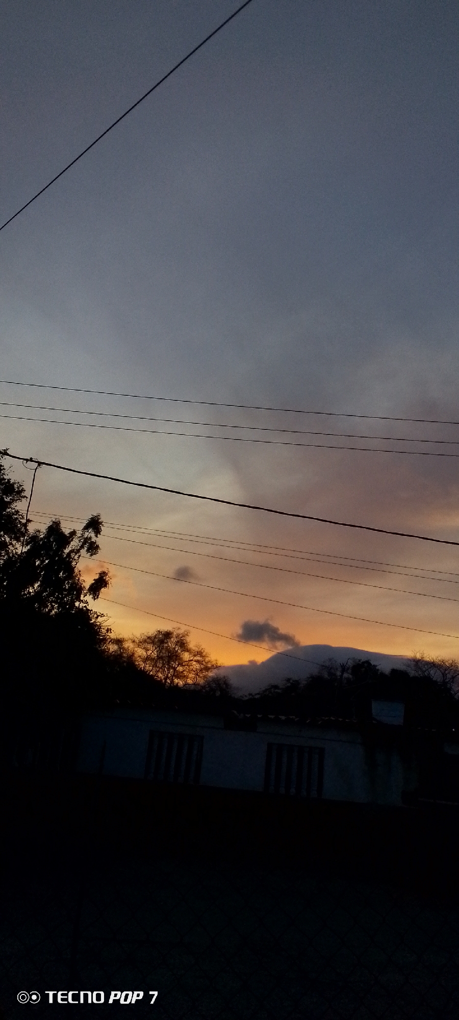 The image shows a sunset in what appears to be Altagracia, Venezuela, based on the city identifier "TECNO POP 7" potentially associated with a mobile device used in the region. The sky transitions from a dark grey-blue at the top to an orange hue closer to the horizon. Silhouetted against the sunset are trees, foliage, and what may be a small building with windows. Power lines cross the upper portion of the frame. A dark shape resembling a mountain is present in the distance. The foreground is mostly dark, obscuring details of any ground or objects at the bottom.