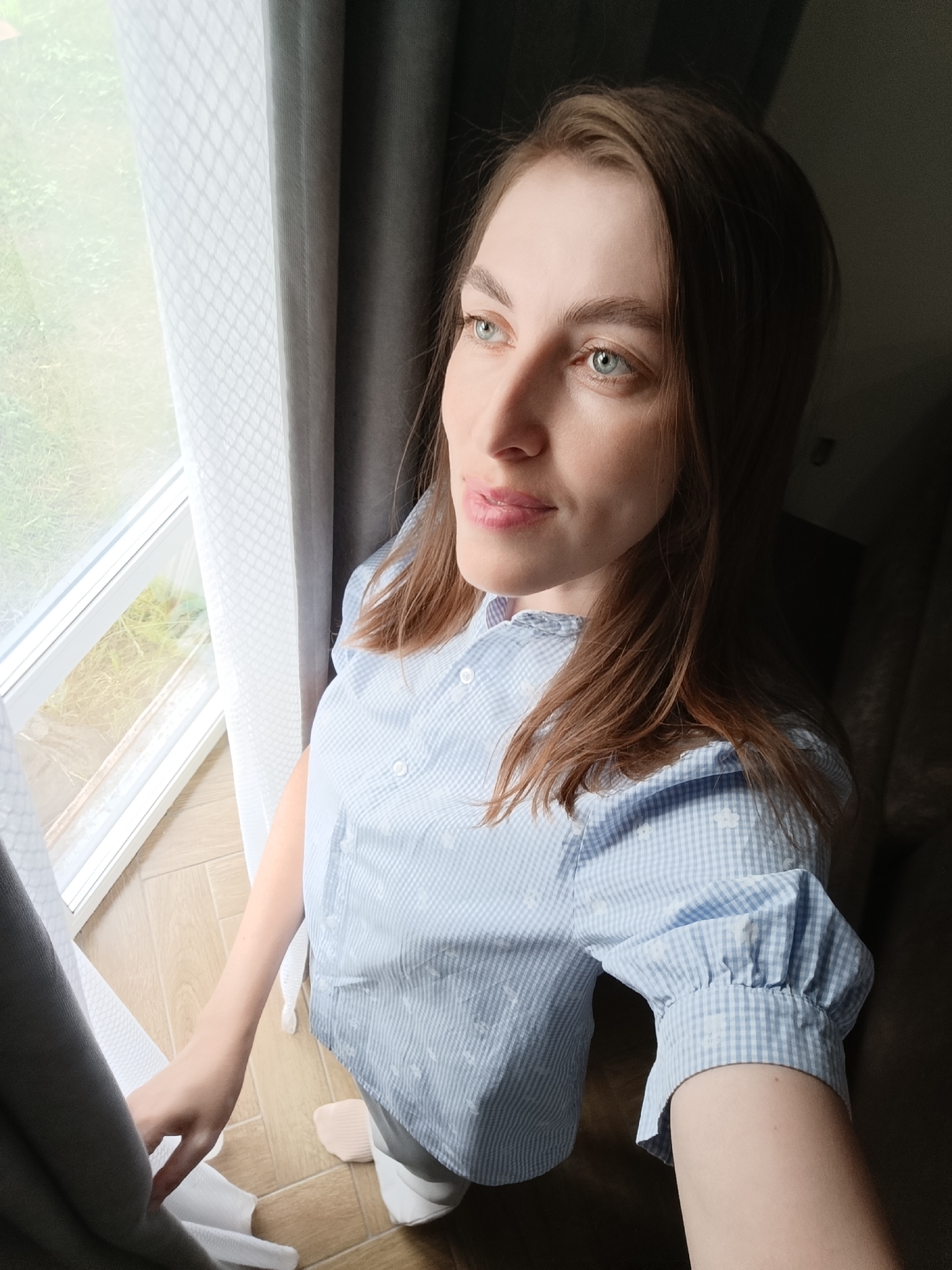 A woman with light brown hair and green eyes is looking out of a window. She is wearing a blue and white checkered shirt. The window has white sheer curtains and a gray curtain next to it. Outside the window, green vegetation is visible. The photo appears to be taken indoors, with a dark area suggesting a room or corner. The location is Chaikovsky, Russia.