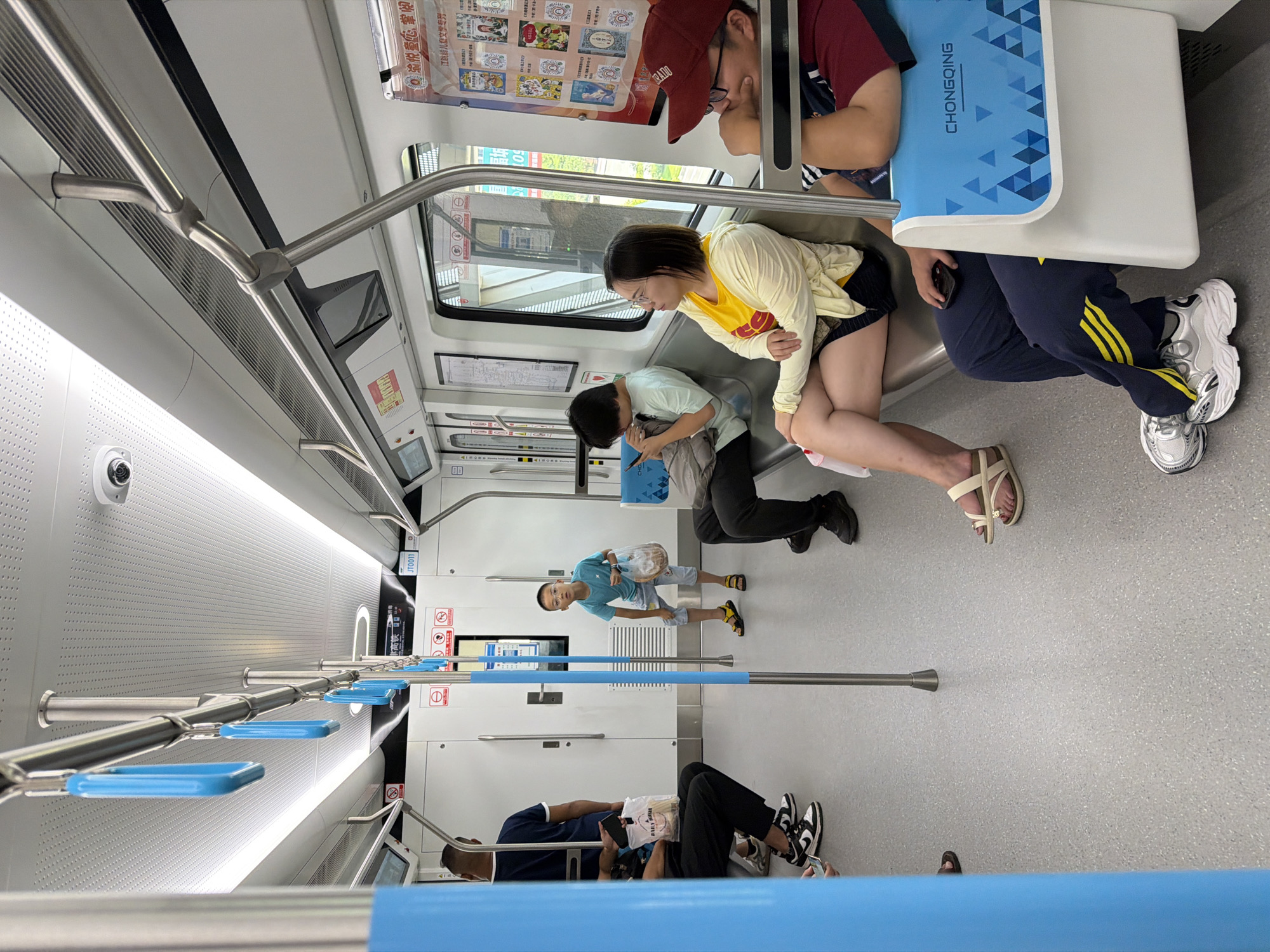 This image shows the interior of a subway car in Chongqing, China, specifically on the Jiangjin line. The car is clean and modern, with white walls and ceiling, and gray flooring. Blue and silver handrails are visible throughout the car. Several passengers are seated and standing. On the left, a person in a blue shirt is standing, facing away from the camera. In the middle of the car, a young boy in a blue t-shirt and shorts is standing, holding a plastic bag. To his right, a woman wearing a yellow shirt and black shorts is seated, looking to the left. In the foreground, a person wearing a red cap and glasses is seated, looking to the right. The flooring is a speckled gray material, and the seats appear to be a light gray color. Posters with colorful graphics are displayed on the right wall of the car. The overall impression is of a functional and well-maintained public transportation environment.