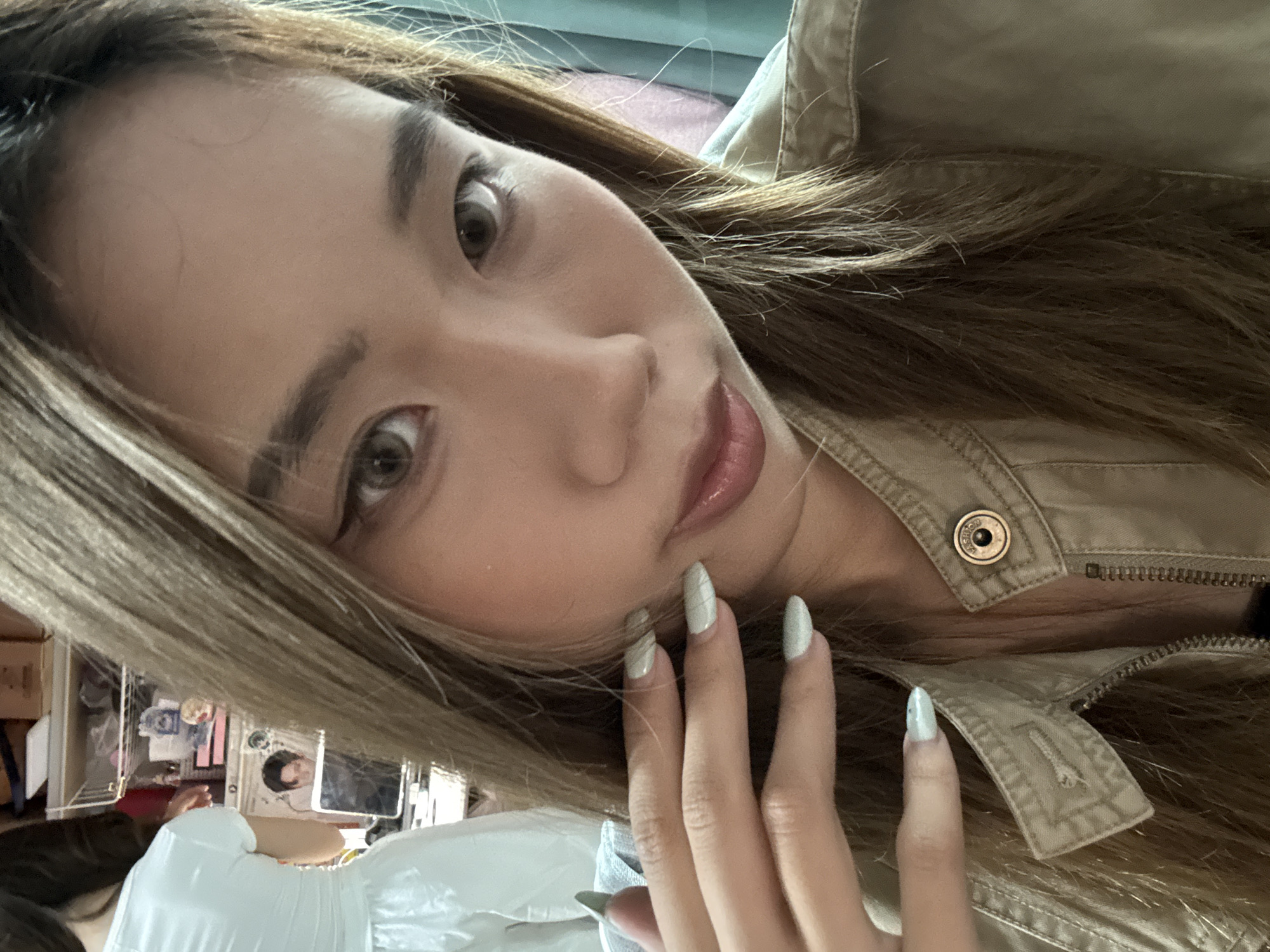A young East Asian woman is taking a selfie. She has long, light brown hair, light brown eyes, and is wearing makeup. Her left hand is raised to her face, with light green fingernails visible. She is wearing a light brown collared jacket with a zipper and a button. In the background, a person in a white dress and a laptop are visible. The image appears to be taken indoors. The location is not discernable from the image.