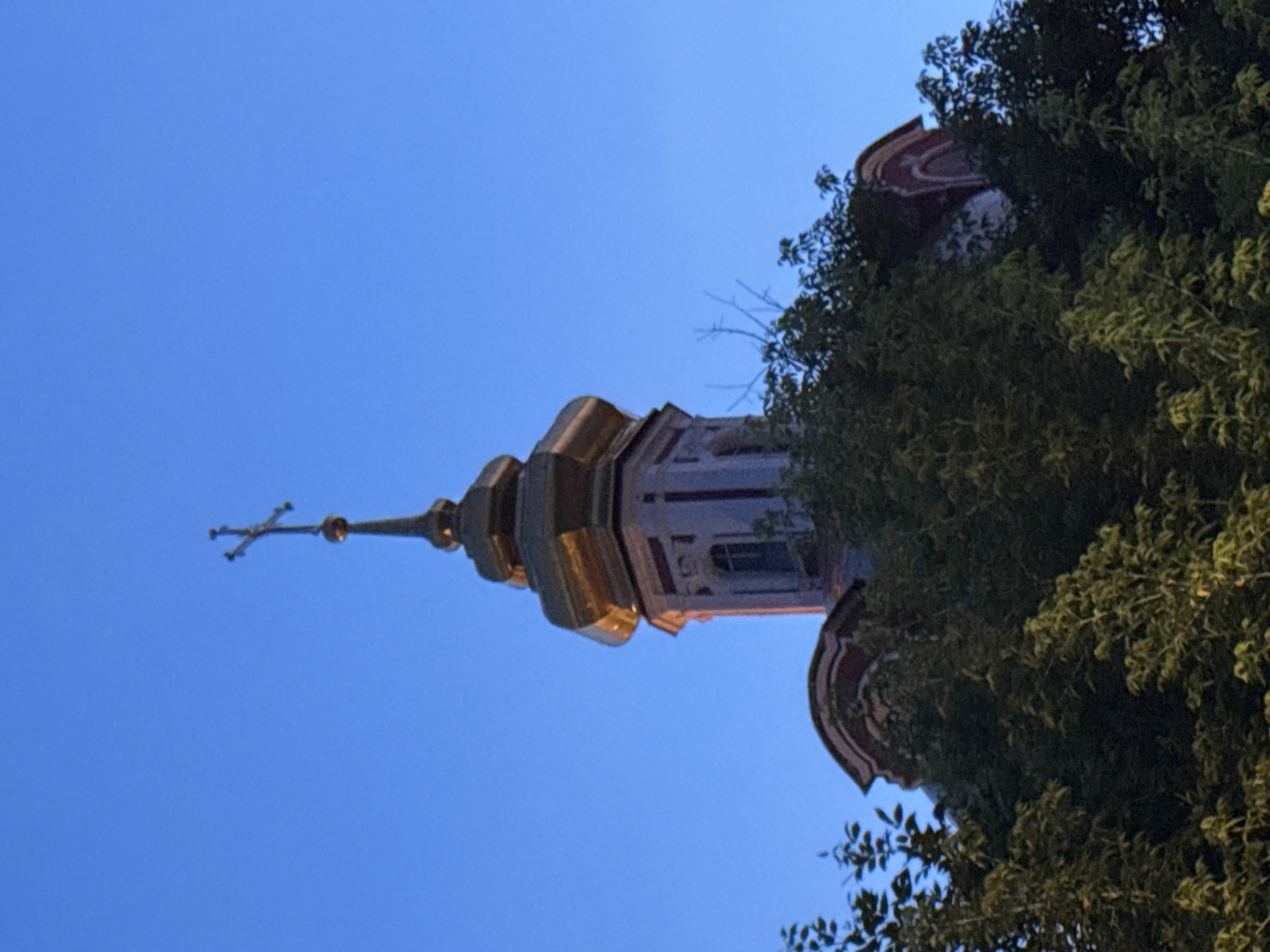 The image shows the dome and cross of a church against a clear blue sky. The dome is gilded and octagonal, with decorative trim. Below the dome is a section of the church tower, which appears to be made of stone with red trim. The church is partially obscured by lush green trees in the foreground. The lighting suggests it is either early morning or late evening. The scene depicts a religious building in an urban environment, with vegetation contributing to the overall aesthetic. This church is identified as the Church of the Ascension of the Lord in Kolomenskoye, Moscow, Russia.