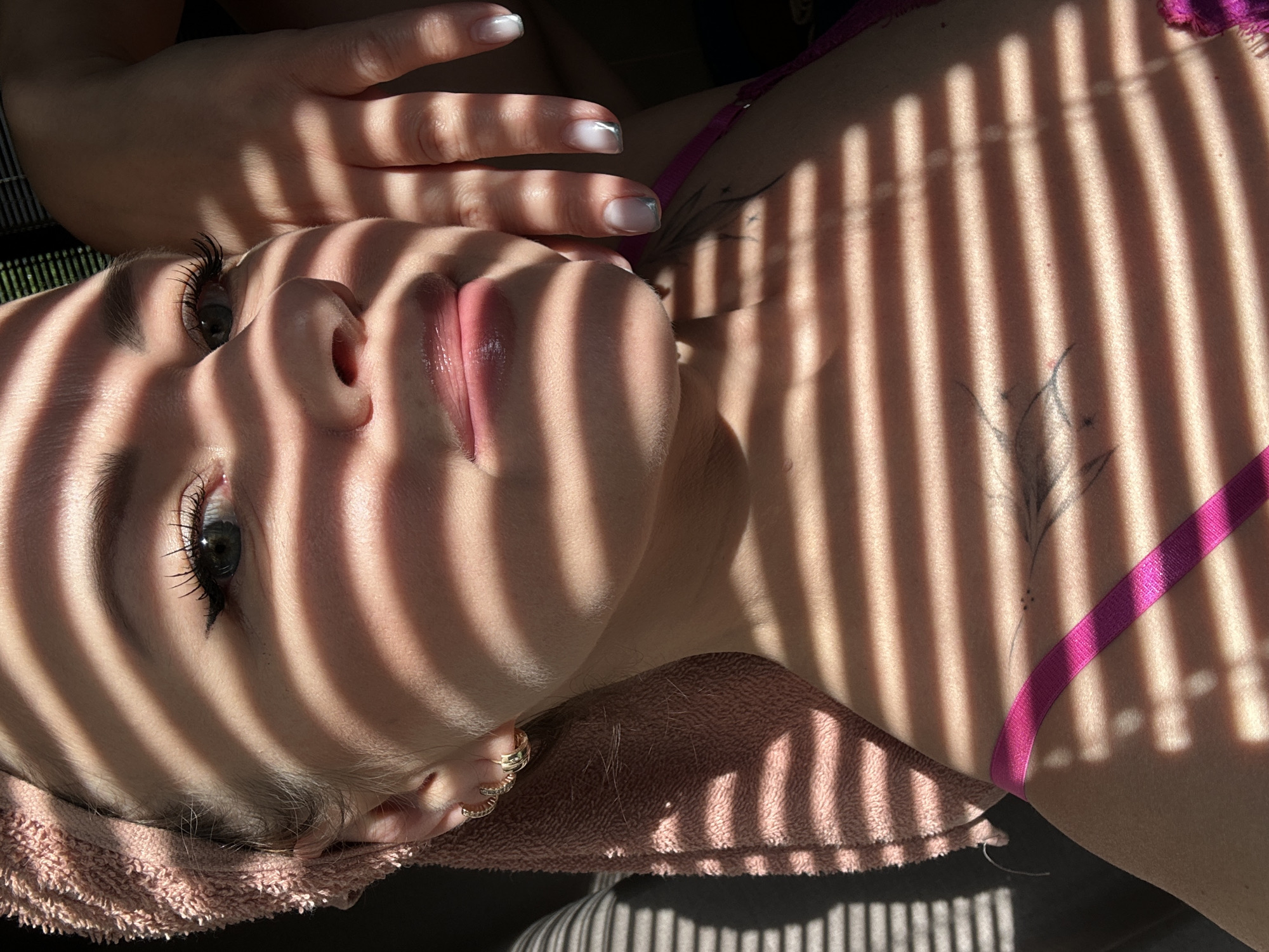 A close-up, high-angle shot captures a person, likely a woman, reclining, bathed in strong, parallel stripes of sunlight and shadow. Her face is prominently featured, with her right eye open and looking upwards, revealing a dark iris and accentuated eyelashes. Her lips are slightly parted. Her head rests on a soft, pink, possibly terry cloth, towel.
Her hand is visible near her face, showcasing fingernails with a French manicure featuring silver tips. A gold hoop earring adorns her left earlobe. She wears a bright pink or magenta strap, likely from a bra or swimsuit, over her right shoulder. A subtle tattoo, appearing as a stylized floral or organic design, is visible on her right upper torso, partially obscured by the shadow stripes.
The distinct pattern of light and shadow suggests bright sunlight entering an indoor or semi-enclosed space, likely filtering through blinds or a slatted structure. This indicates it is daytime, probably late morning or afternoon. The background is dark and out of focus. The person's expression appears calm and contemplative. No text is visible in the image. The scene is located in Chaikovsky, Russia.