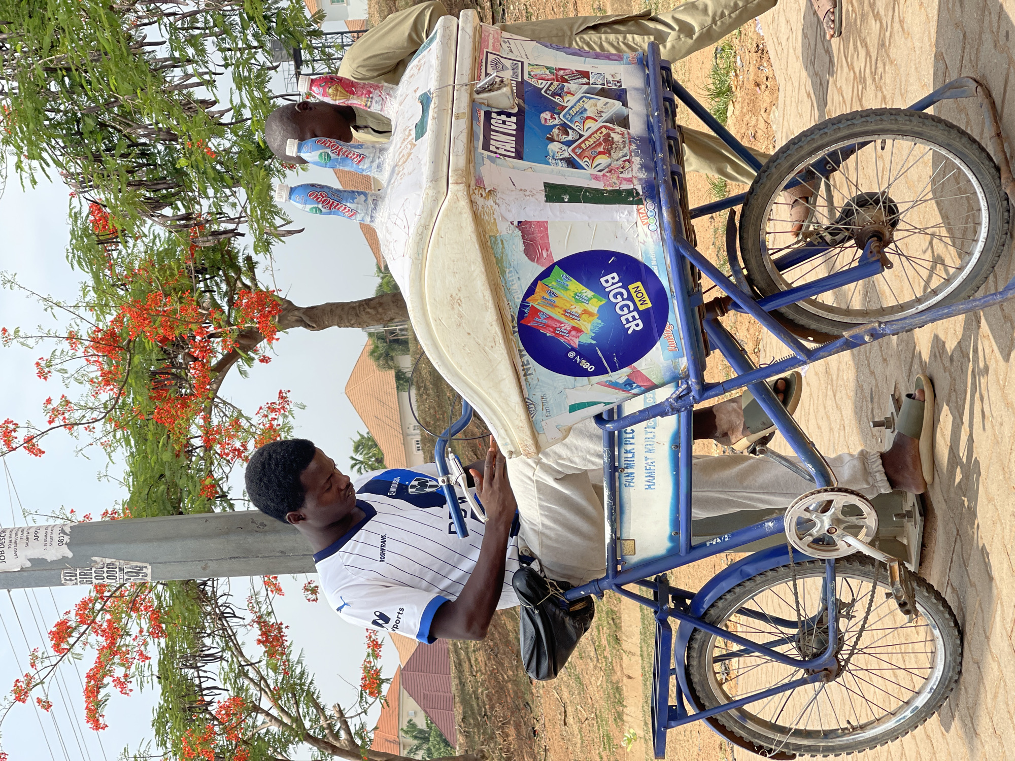 A man is sitting on a blue tricycle with an ice cream cooler in Kuchingoro, Nigeria. The cooler is white and covered in advertisements for Fan Milk products and the cooler has several yogurt drinks displayed on top. The man is wearing a white soccer jersey with blue trim and a black bag slung across his body. The tricycle is parked on a paved surface, and there is another man standing on the other side of the cooler. In the background, there are trees with red flowers and buildings.