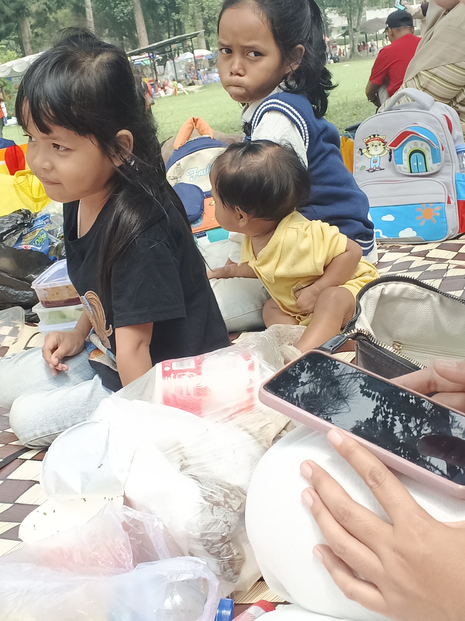 In the city of Perbawati, Indonesia, a group of children and adults are gathered for an outdoor picnic. The foreground is occupied by several children, with two girls and one baby prominently featured. One girl, on the left, wears a black t-shirt and light blue jeans, looking off to the side. Beside her, a younger girl in a blue and white vest and white shirt faces the camera with a pouting expression. A baby in a yellow onesie is nestled between them, facing away from the camera.
Surrounding the children are picnic essentials: a checkered mat, various food containers, plastic bags filled with items, and a child's backpack with colorful cartoon characters. A person's hands hold a pink smartphone, with reflections of trees and sky visible on its screen. In the background, other people and picnic setups are visible, along with trees and a tent structure, suggesting a park or recreational area. The overall atmosphere appears relaxed and familial, indicative of a casual outdoor gathering.
