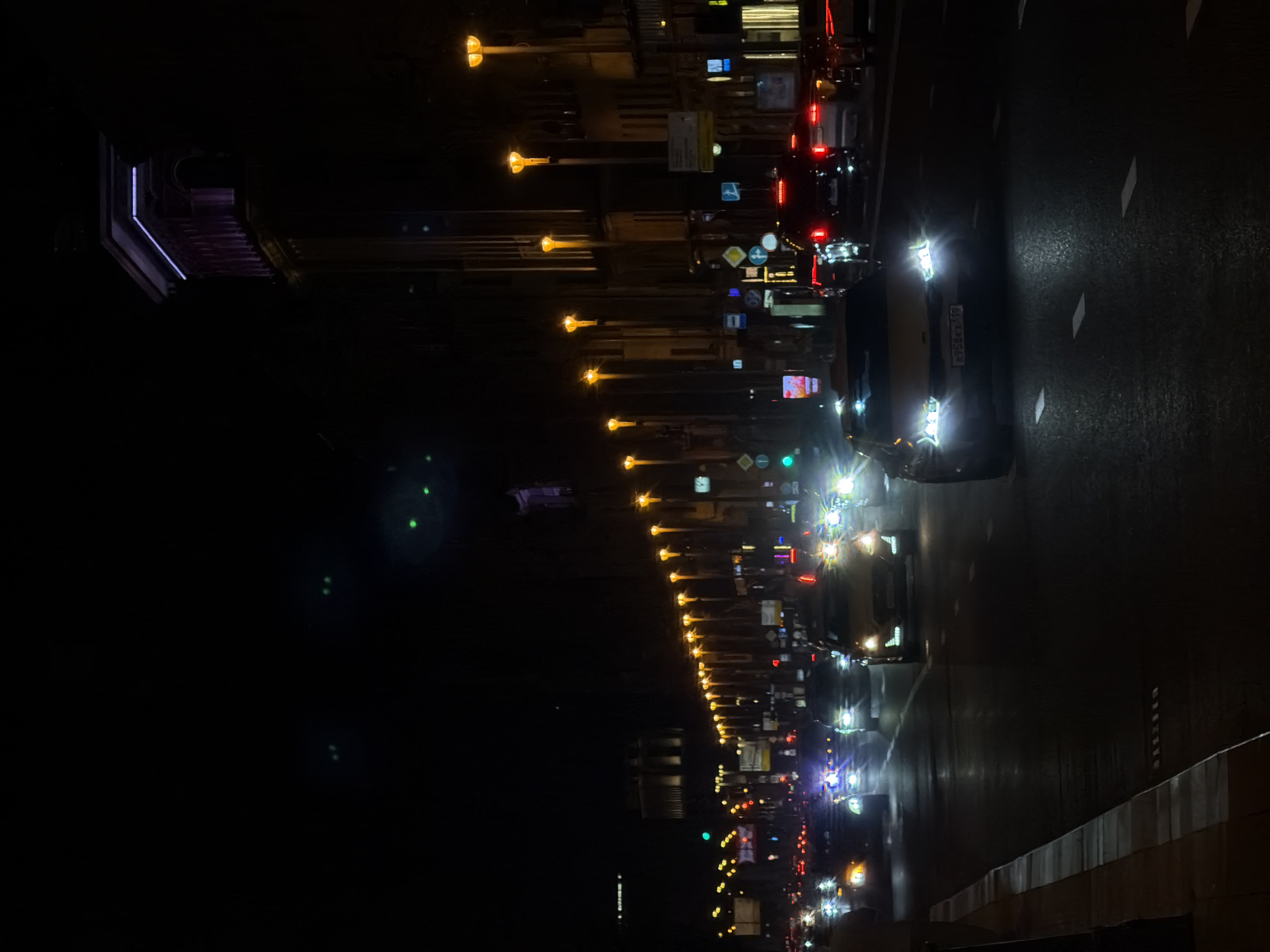A nighttime street scene in Moscow, Russia, shows a road lined with streetlights. Cars with headlights and taillights are visible on the right side of the image. The scene appears to be taken from an elevated position, with a bridge or structure visible to the left. Street signs and other urban infrastructure elements are also present.