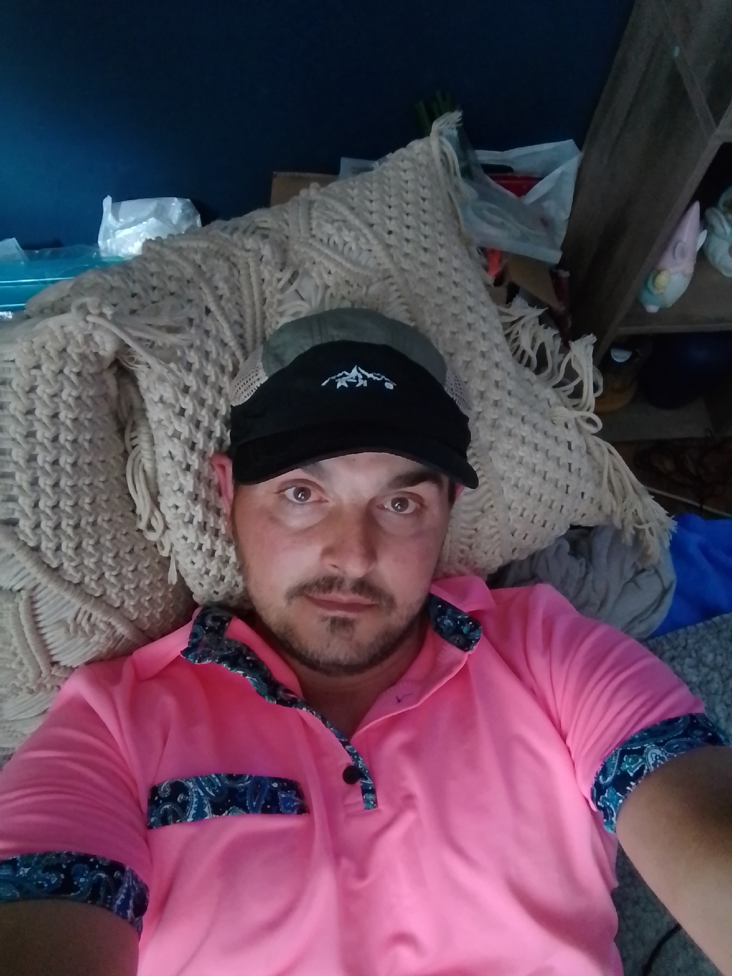 A man wearing a pink polo shirt with blue paisley accents and a black baseball cap with a mountain logo is lying on a textured, cream-colored blanket. The man is looking directly at the camera. The background is dark and out of focus, with some shelving and objects visible to the right. The overall scene appears to be indoors, possibly in a living room or bedroom. There is no information in the image to suggest a specific location in Wedel, Germany.