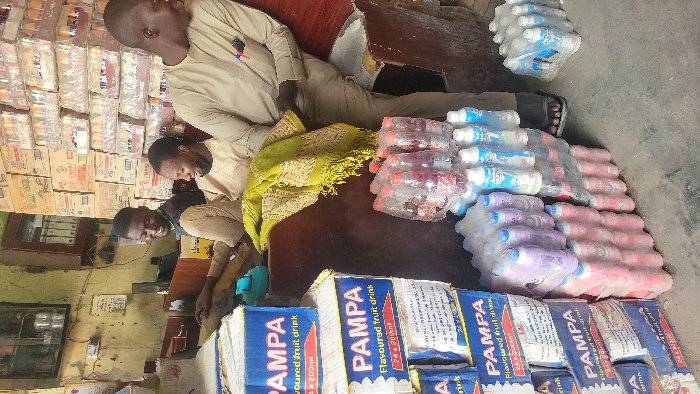 The image depicts an indoor retail space in Fagge A, Nigeria, characterized by stacked merchandise and three adult males interacting. On the left foreground, multiple blue and white cardboard boxes labeled "PAMPA Flavoured fruit drink, 24 x 200ml" are stacked vertically. To the right of these boxes, various plastic-wrapped bundles of bottled beverages are arranged on a concrete floor, including clear bottles with red contents, purple/pink bottles with blue caps, and smaller clear bottles with white caps.
Behind the beverage stacks, two men are seated or leaning behind a dark wooden counter. The man on the left wears a black short-sleeved shirt and light brown trousers, smiling and looking towards the right. The man in the middle wears a light brown traditional top and trousers, with a black face mask pulled below his chin, also smiling and looking right. A green patterned fabric with fringe is draped over the counter between the middle man and the man on the right.
The third man stands or leans on the right side of the counter, wearing a light brown traditional top with pens clipped to his chest pocket, dark trousers, and black flip-flops. He is smiling and looking towards the left, appearing to interact with the other two.
The background features a light-colored wall with some visible wear. On the left wall section, a dark framed item, an electrical outlet, and exposed wiring are present. To the right, a wooden cabinet with glass doors holds documents or files. Further right, the wall is extensively covered by stacks of cardboard boxes containing various products, suggesting a storage or sales area for bulk items. The overall lighting is consistent with an indoor setting during daylight hours.