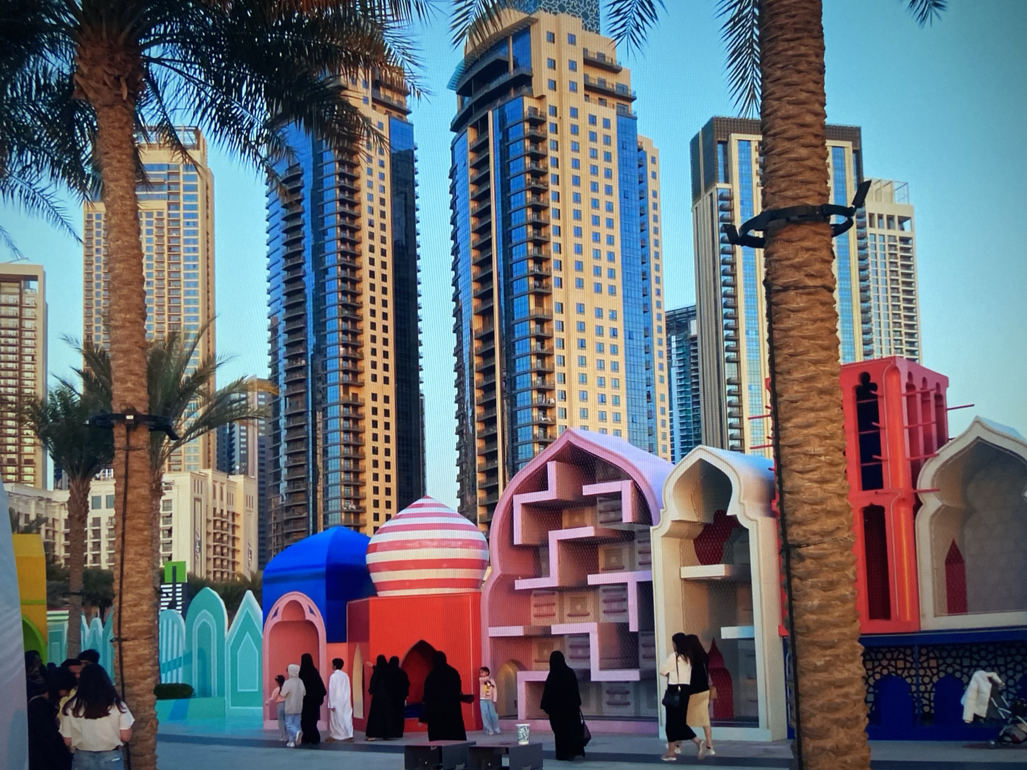 The image depicts a public space in an urban setting, with a mix of modern architecture and culturally-themed structures. Several high-rise buildings form the backdrop, displaying a modern architectural style with glass and concrete exteriors. Palm trees are scattered throughout the foreground, framing the scene.
In the foreground, there are colorful, stylized architectural structures that appear to be inspired by traditional Middle Eastern designs. These structures feature a mix of pastel colors and geometric patterns. People are present in the scene, some dressed in traditional attire, suggesting a cultural event or attraction. Interactions among individuals are observed, with some appearing to be tourists taking photos or families enjoying the space. The overall atmosphere is vibrant and inviting, blending modern urbanism with cultural expression.