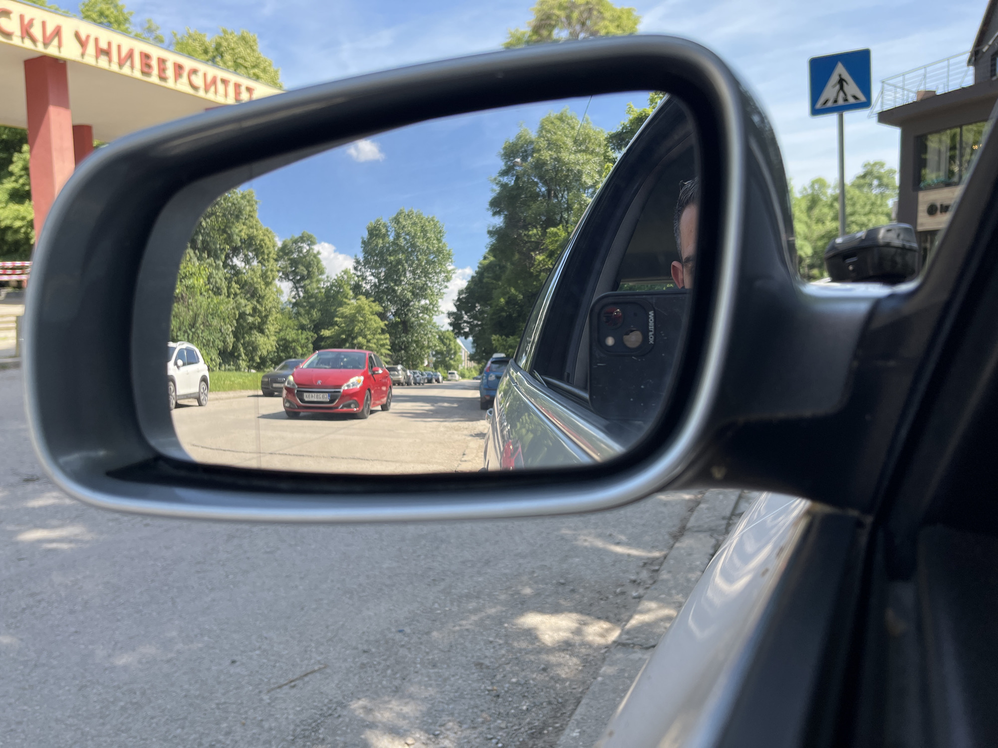 The image captures a reflection in a car's side mirror, showing a street scene in Sofia, Bulgaria. In the reflection, a bright red car is prominently visible, stopped in traffic. Behind it, several other vehicles are lined up on the asphalt road. Trees with lush green foliage line the street, and the sky above is a clear blue with scattered white clouds. To the left of the reflection, a large sign with Cyrillic lettering is partially visible, appearing to be the entrance to a university. A pedestrian crossing sign with a blue background and a white silhouette of a person is also visible in the reflection. The car's interior is visible in the foreground of the reflection, including a portion of the driver's face and a smartphone mounted on the dashboard. The exterior of the car and the road outside the reflection are also visible in the main image.