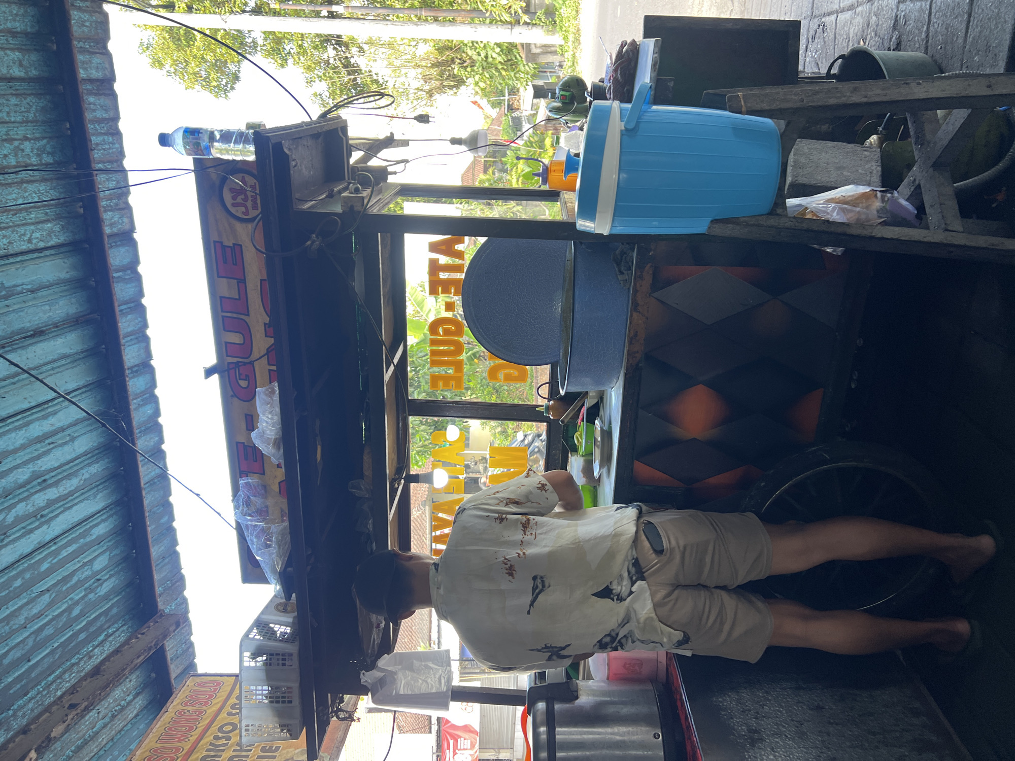 In the city of Payangan, Indonesia, a man stands behind a food cart under a blue corrugated metal roof. The man, wearing a light-colored short-sleeved shirt with a bird motif and khaki shorts, is facing away from the camera, appearing to prepare food. The food cart is open, with a glass front displaying food items and signage. Prominently displayed are signs with text, including "AKSO SOLO" and "GULE-GULE." A large blue cooler and a white plastic dispenser are visible on the right side of the cart, along with various containers and cooking utensils. In the background, a street with trees and buildings can be seen. The scene suggests a small, local food vendor operating on a street in Payangan.