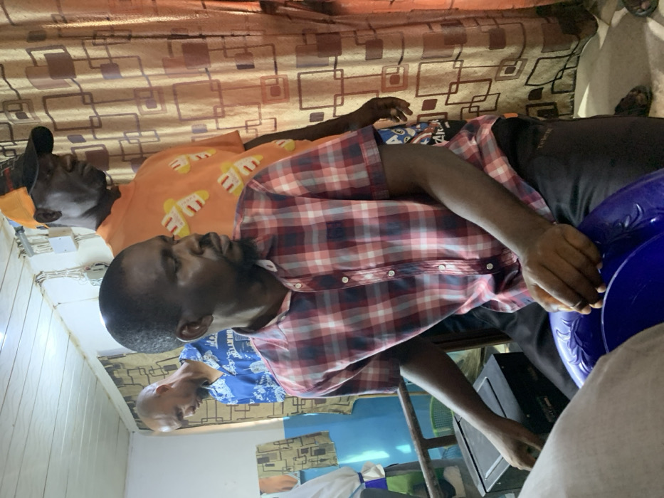 Within an indoor setting in Okene, Nigeria, three adult males are present.
In the foreground, an adult male with a dark complexion, short dark hair, and a trimmed beard and mustache is seated or crouched, facing slightly to his right. He wears a short-sleeved red, white, and blue plaid shirt and dark trousers. His hands rest on a blue plastic basin with embossed floral patterns.
Behind and to his right stands a second adult male with a dark complexion, wearing an orange baseball cap with a dark crown. He has on a short-sleeved orange t-shirt patterned with multiple yellow and white slipper-like designs. This individual is observed looking downwards and to his right.
Partially visible in the left background is a third adult male with a dark complexion and a bald or closely shaven head. He wears a short-sleeved blue button-up shirt featuring a white cityscape pattern and appears to be looking downwards.
The room features a white ceiling with exposed white electrical wiring and conduit runs. The wall on the left side is painted a light blue color, while a patterned brown curtain with a repeating geometric design covers the wall area to the right. A dark, rectangular electronic device is visible on a surface behind the central figure to his left, and a metal frame, possibly from a bed or bench, is also present.
