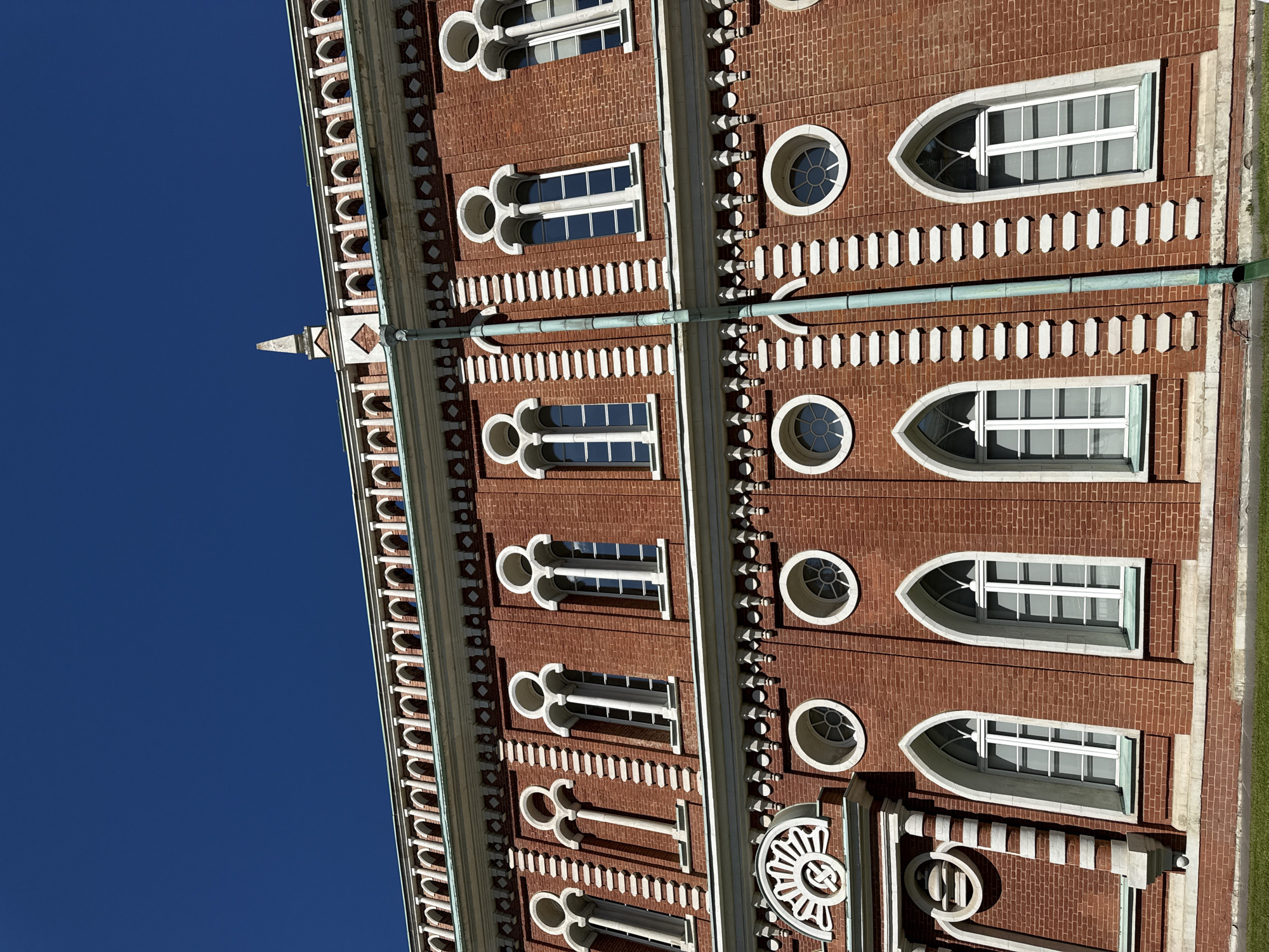 This is a low-angle, full-shot image of the red brick facade of a historic building, likely the Grand Palace in Tsaritsyno Park, Moscow, Russia, against a clear, bright blue sky. The building features intricate architectural details, including a decorative white cornice with repeated arched openings along the top. Below the cornice, there is a band of white diamond-shaped ornamentation. The facade itself is constructed of red bricks and is punctuated by rows of windows.
The upper level of windows are tall and narrow with pointed arches and elaborate white stone tracery. The lower level features a mix of windows: several are arched and tall with white frames, while others are circular with intricate white surrounds. A green downspout runs vertically along the facade, intersecting with some of the decorative elements. The building's base is lined with white stone. In the foreground, a strip of green grass is visible. There are no people or animals in the image. The overall impression is one of grandeur and historical significance.