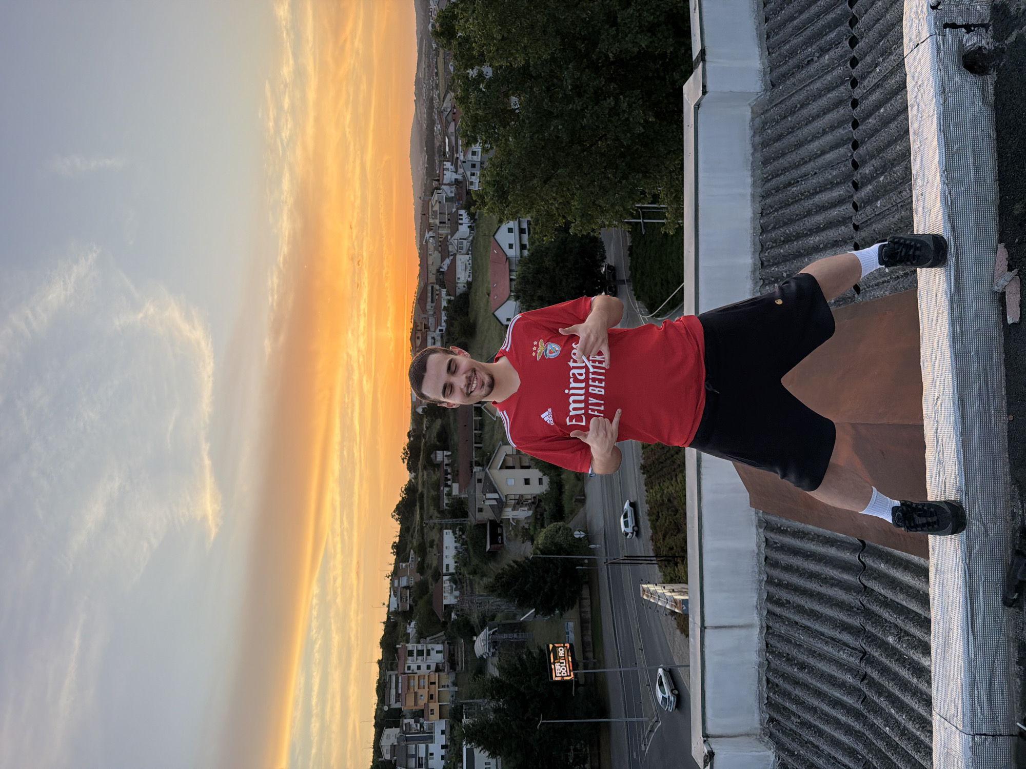 A young man stands on the corrugated metal roof of a building in Bragança, Portugal, against a sunset backdrop. He wears a red Benfica soccer jersey, black shorts, and white socks, and gives two thumbs up. In the background, a townscape with residential buildings stretches towards the horizon, with a road visible with cars and a sign in Portuguese. The sky shows a gradient from blue to orange at the horizon.