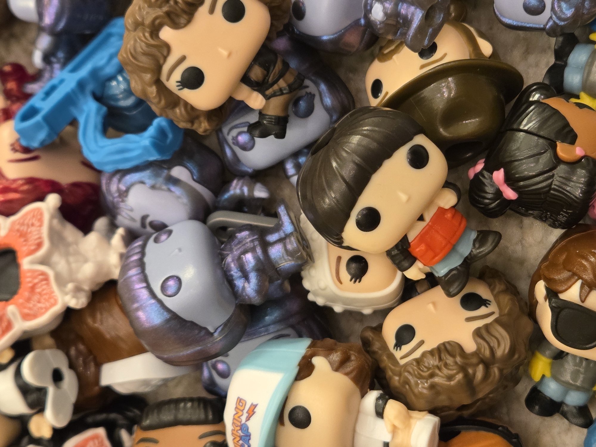 The image presents a close-up view of a collection of Funko Pop figurines. The figurines are jumbled together, with their heads and bodies overlapping. Several characters are visible, including Eleven from Stranger Things, a character wearing a brown hat, and a figure with dark hair. The figurines are made of vinyl, and their features are simplified, with large, round eyes and small mouths. The background is a neutral color. The image does not contain any information that indicates that it was taken in Varna, Bulgaria.