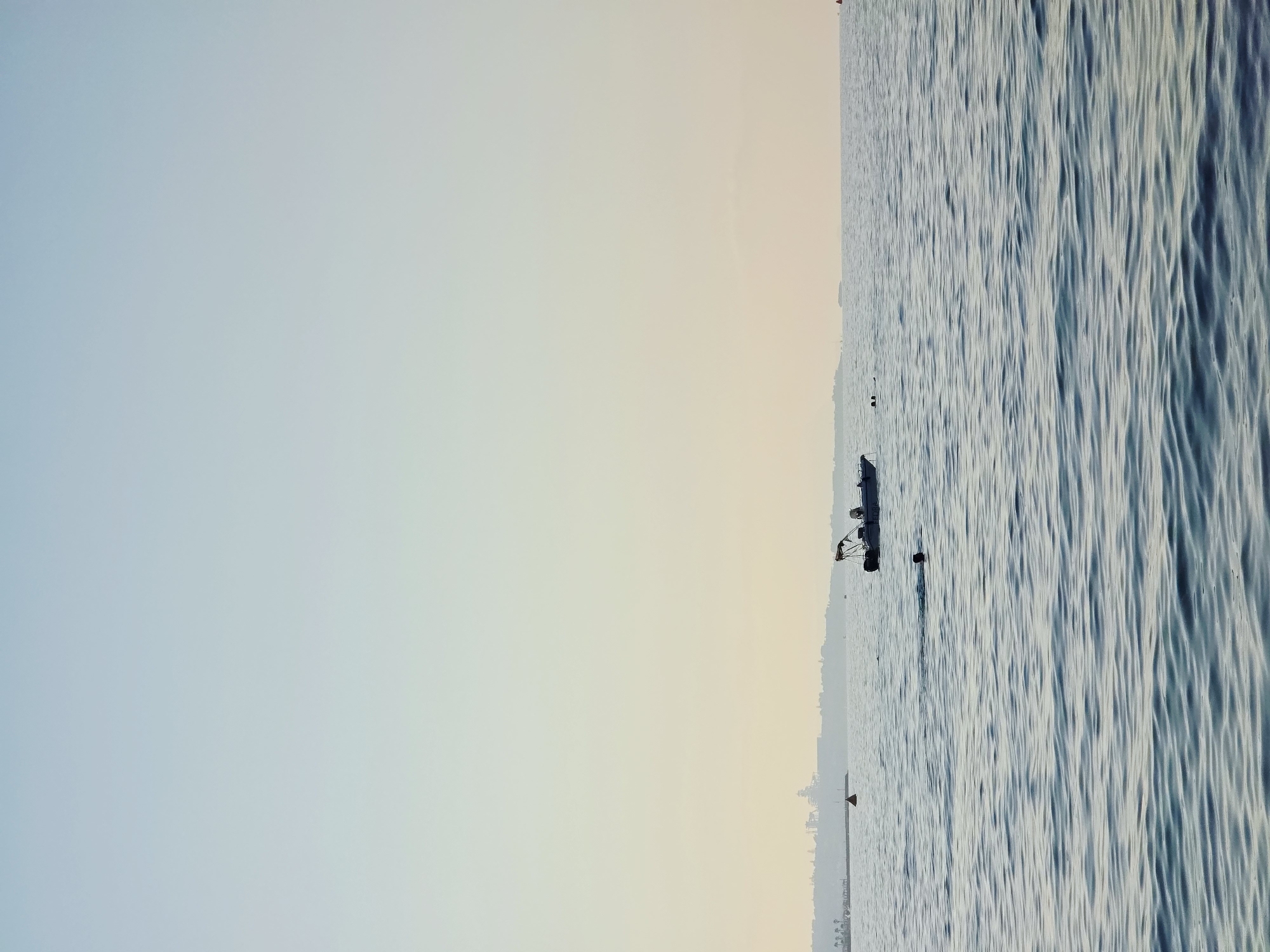 A wide shot captures a serene seascape with a hazy, pale sky transitioning to a warm, pale yellow near the horizon. The water, a deep blue with gentle ripples, fills the lower half of the frame. In the mid-distance, a small white boat with a dark canopy and a few figures aboard is anchored. To the left of the boat, a person is swimming, with only their head visible above the water. Further in the distance, a blurred, silhouetted coastline with buildings and palm trees is visible. A few buoys are scattered across the water. The image evokes a sense of tranquility and a leisurely day at sea, possibly in the Mediterranean.
