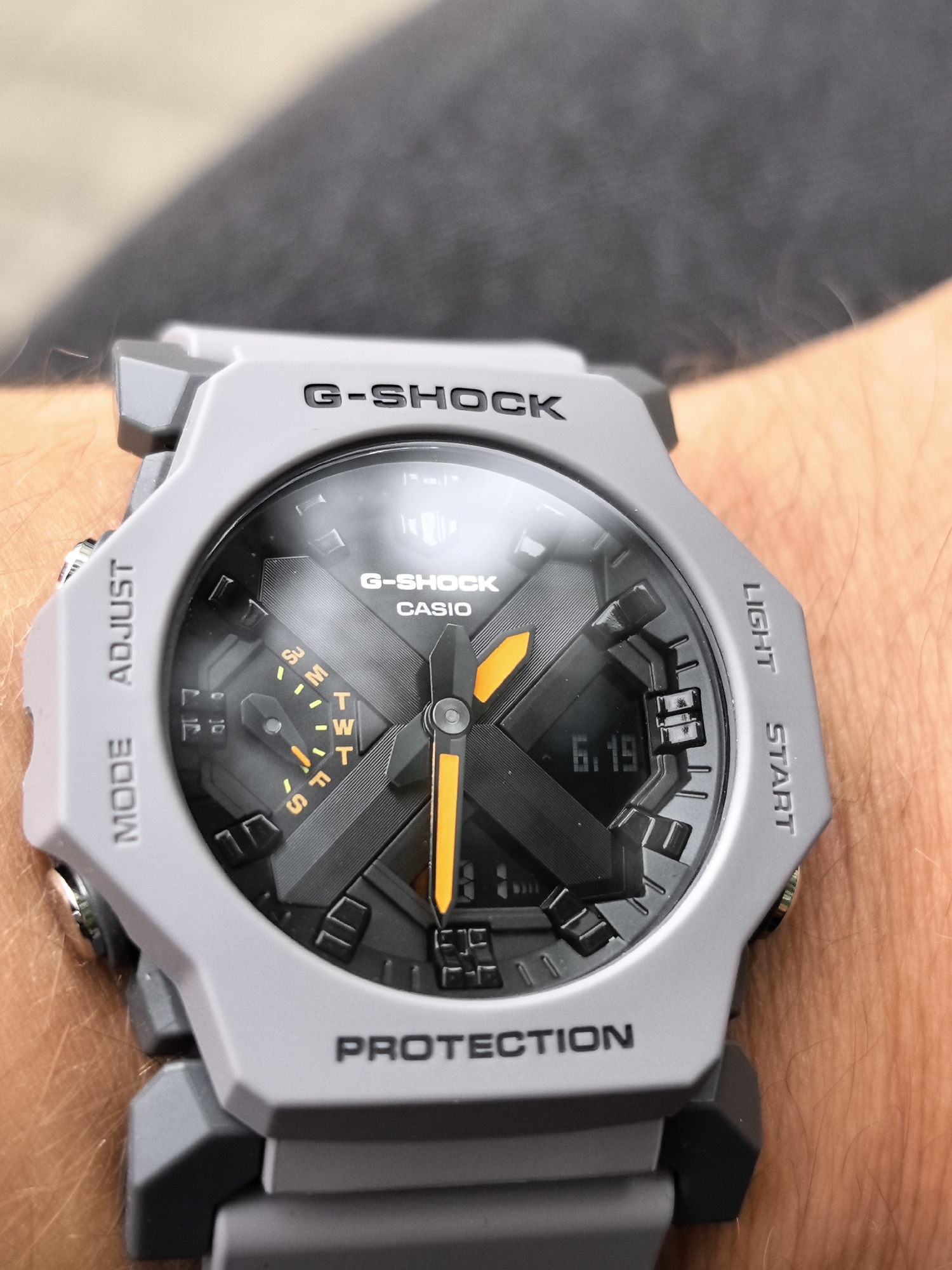 A close-up shot shows a gray G-Shock watch on a person's wrist. The watch face is black with orange accents, and the time displayed is 6:19. The watchband is also gray. The background is blurred and appears to be an outdoor setting.