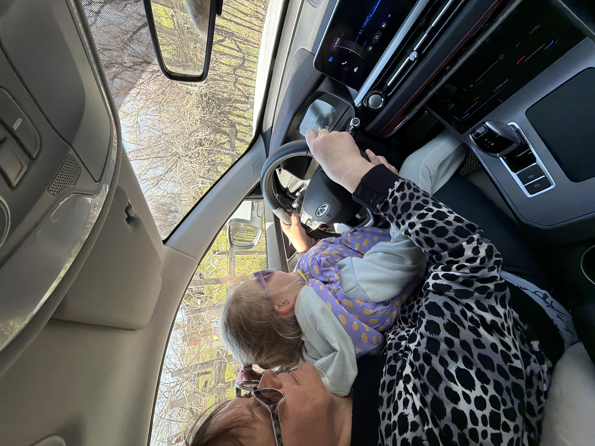 A woman is driving a car with a young child sitting in her lap, possibly in the passenger seat. The car's dashboard, steering wheel, and gear shift are visible. The view through the windshield shows trees. The car is driving near Kamyshin, Russia.